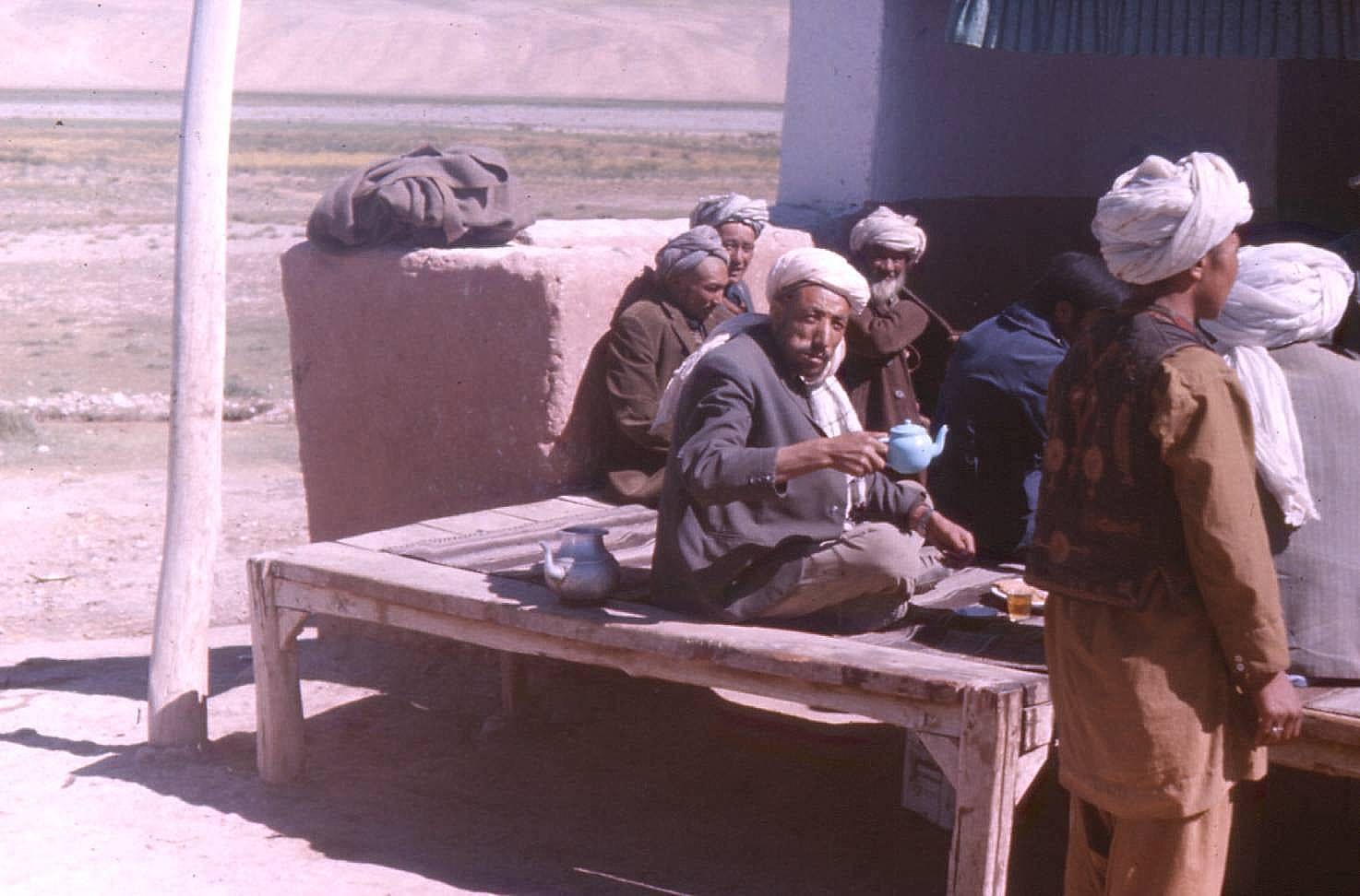 Several men in white and grey turbans sit on a raised wooden platform outside a teahouse, one lifting a small blue cup, with a metal teapot beside him