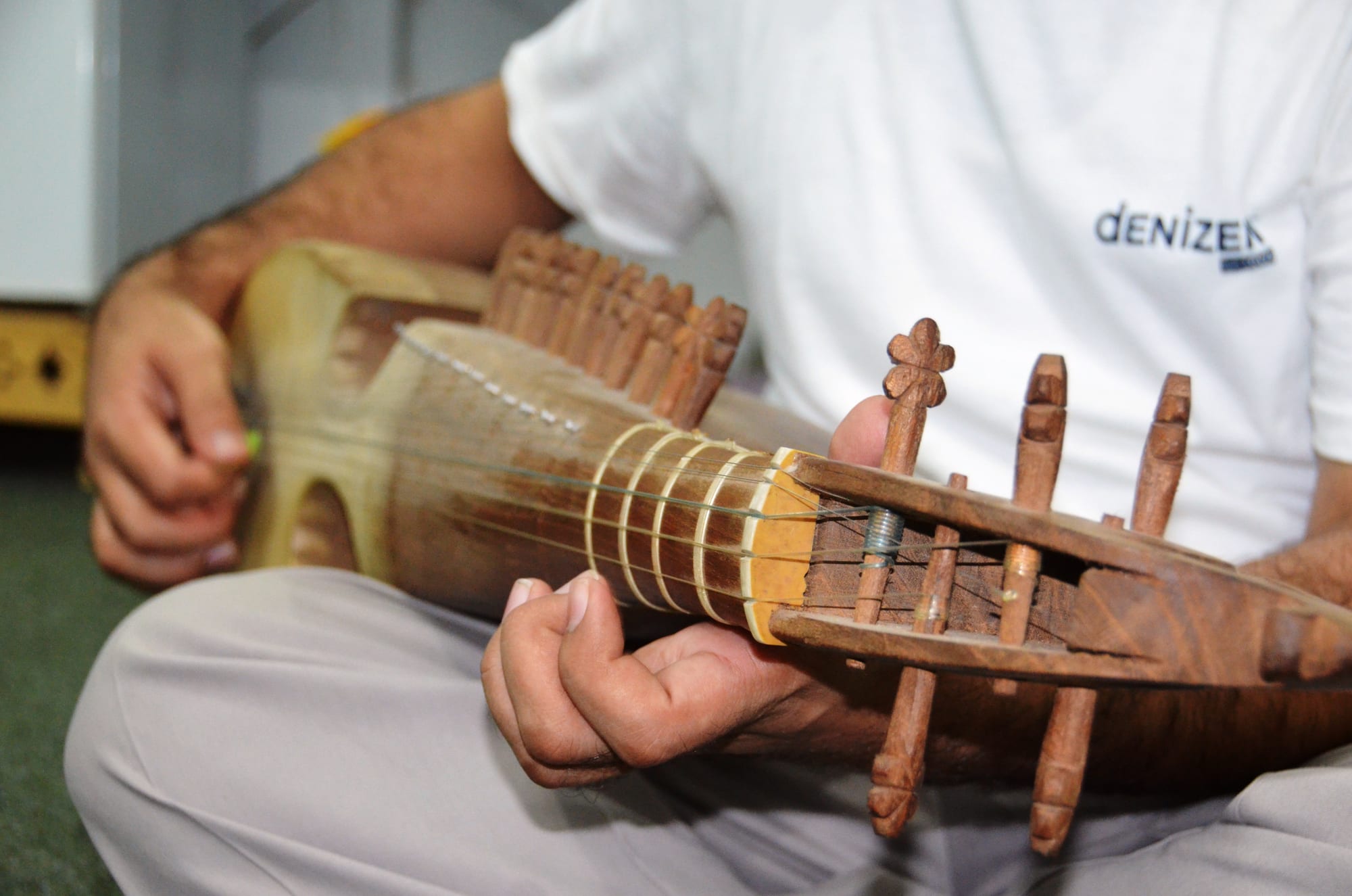 Close-up of a musician's hands on a rubab, one hand fretting the neck and the other resting on the wooden body, with carved wooden tuning pegs in the foreground