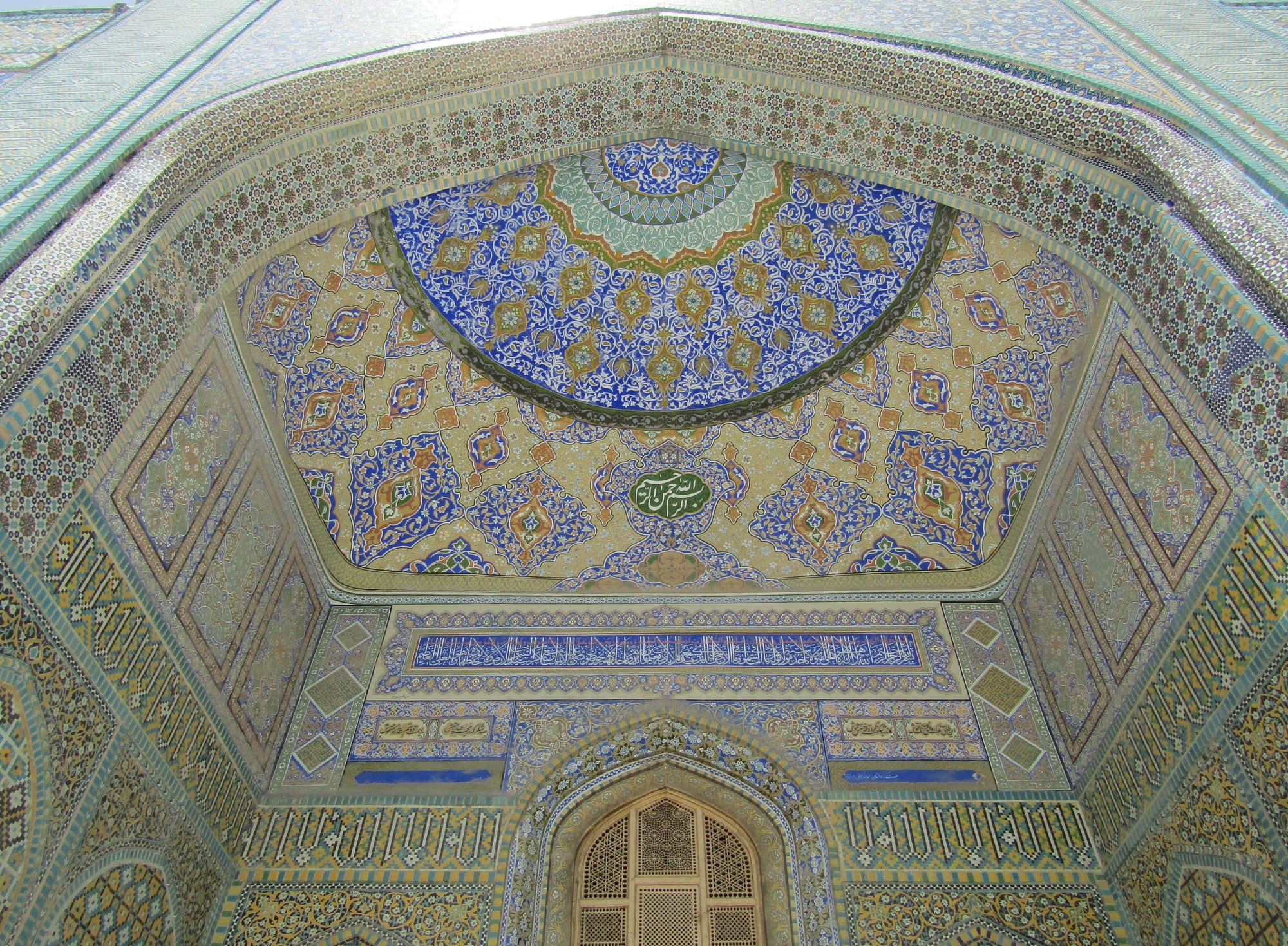 Looking up into a tiled entrance vault covered in blue, turquoise and gold geometric and floral patterns, with a band of white calligraphy on blue above a latticed wooden window
