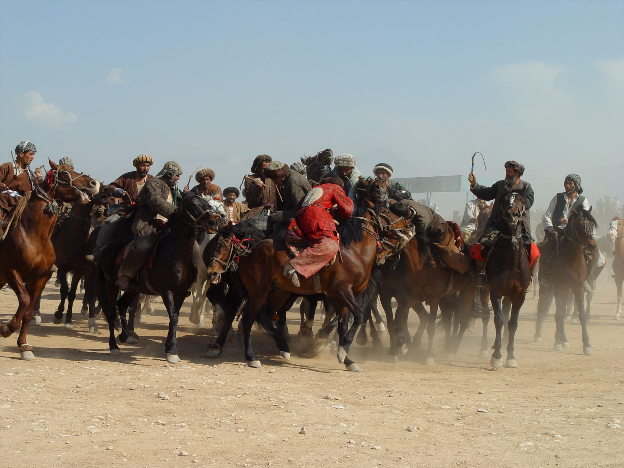 A tight pack of horsemen in turbans and padded coats jostle together on a dusty field, one rider in red leaning low from his saddle, another raising a whip