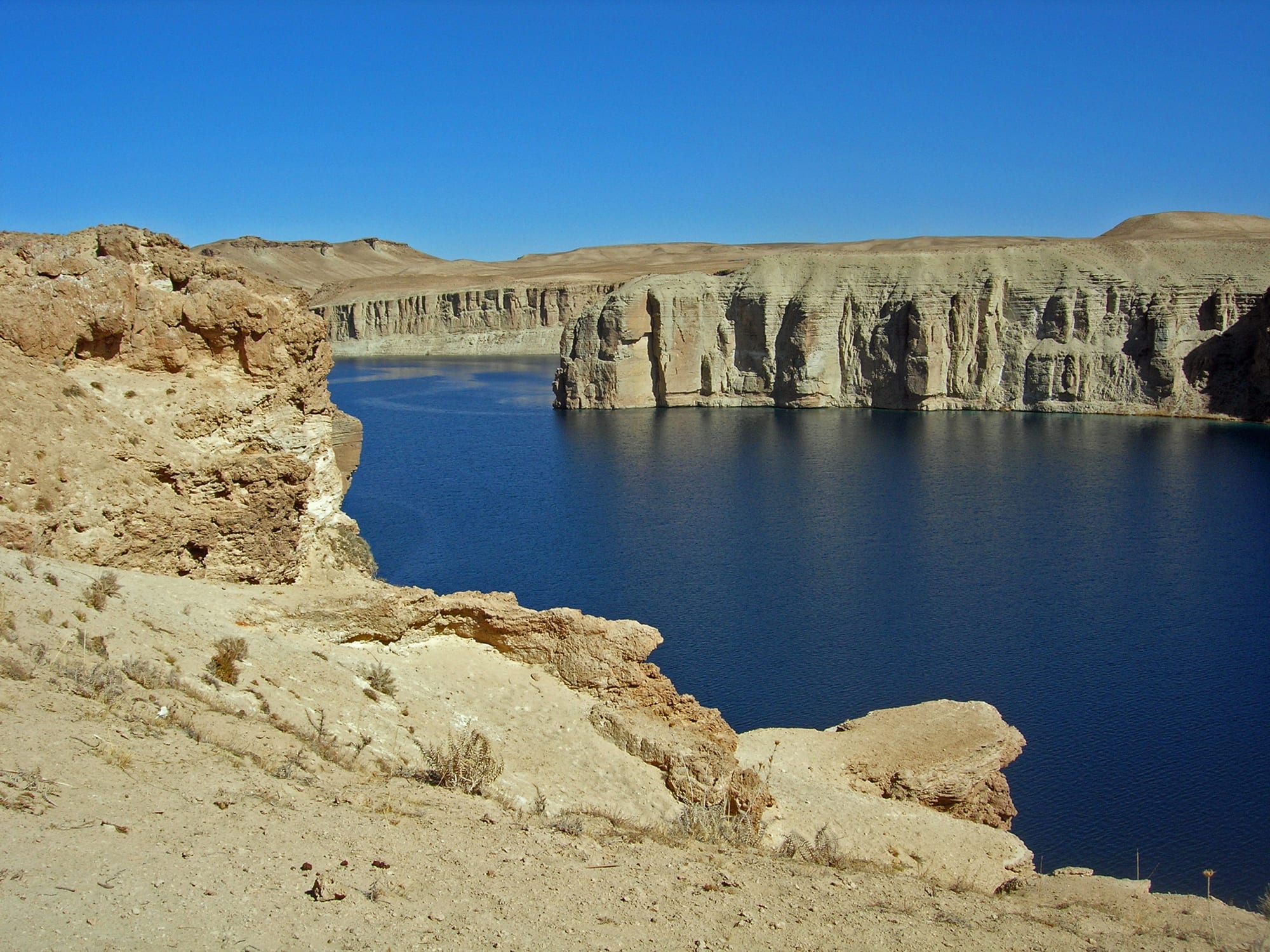 A vivid deep-blue lake held back by a pale natural stone wall, with dry red cliffs behind