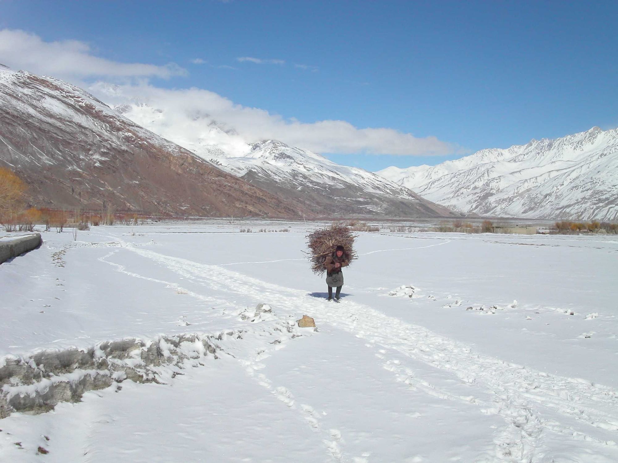 A broad high-altitude valley under light snow, with a lone figure gathering firewood below bare peaks