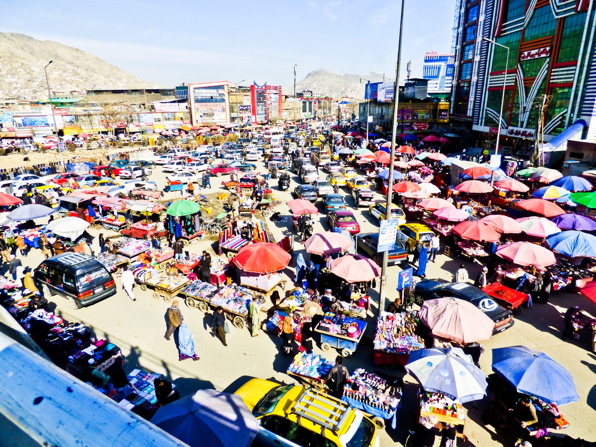 An aerial view of a crowded market square in Kabul with cars, umbrella-covered stalls and pedestrians