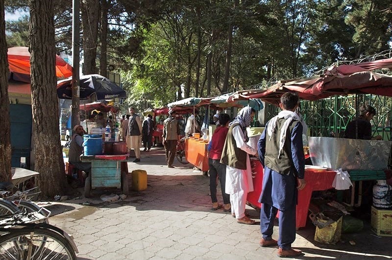 Street food stalls under a tree-lined avenue in Kabul, with several people buying food and bicycles parked nearby