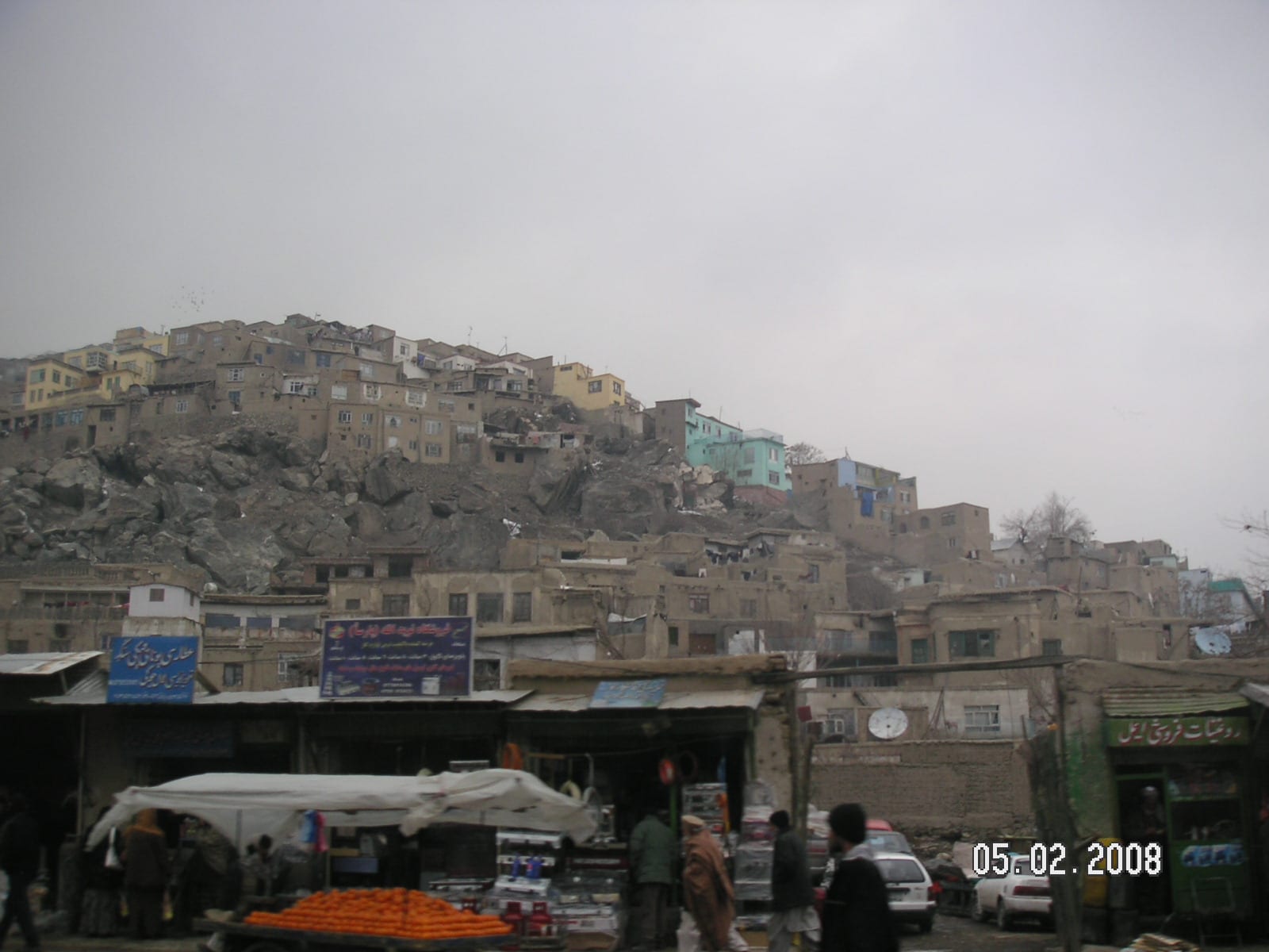 Densely packed houses climbing a rocky hillside above a market street in Kabul