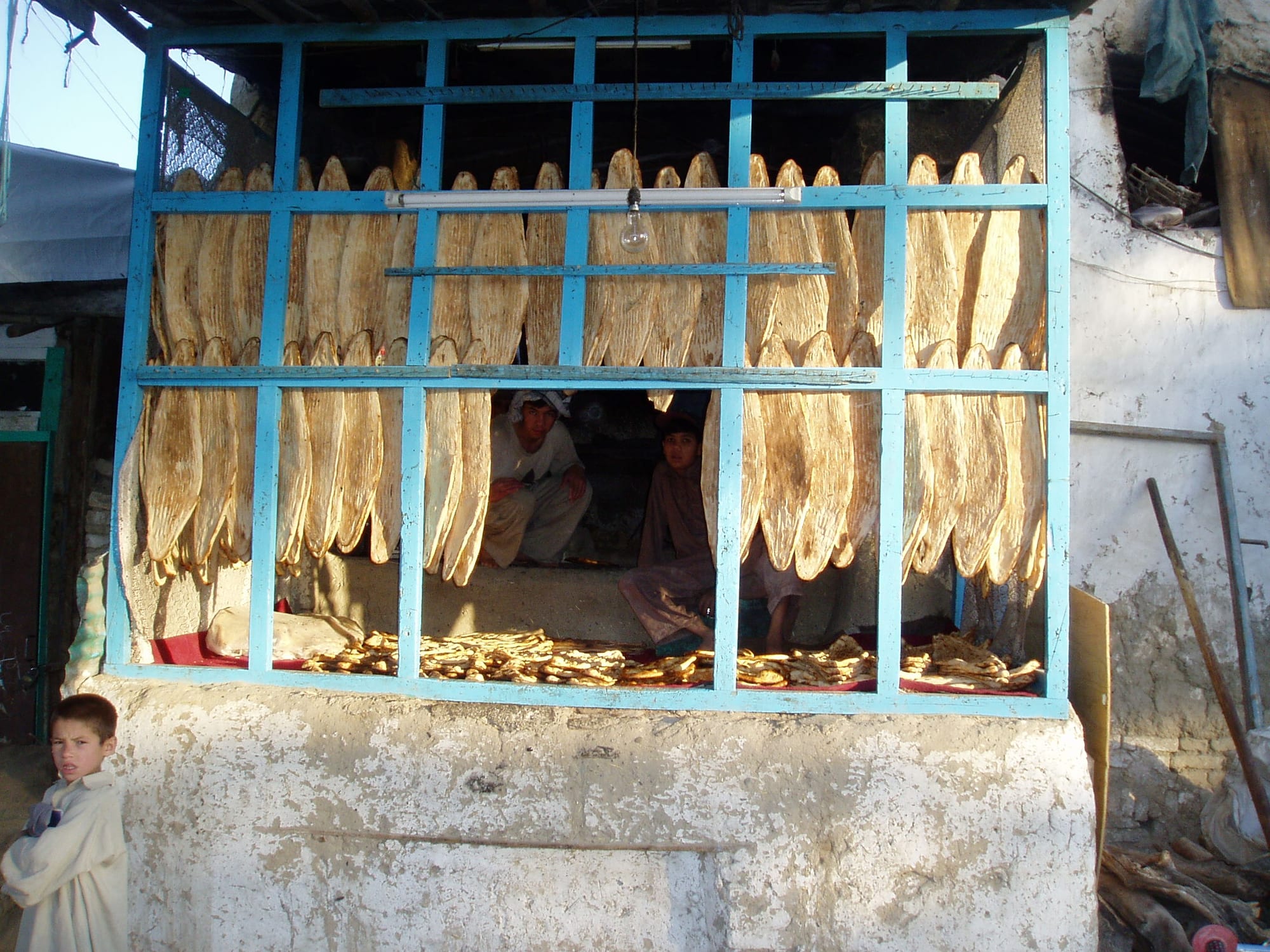 A bakery stall with long paddle-shaped flatbreads hung in rows on a blue lattice front, two bakers visible inside