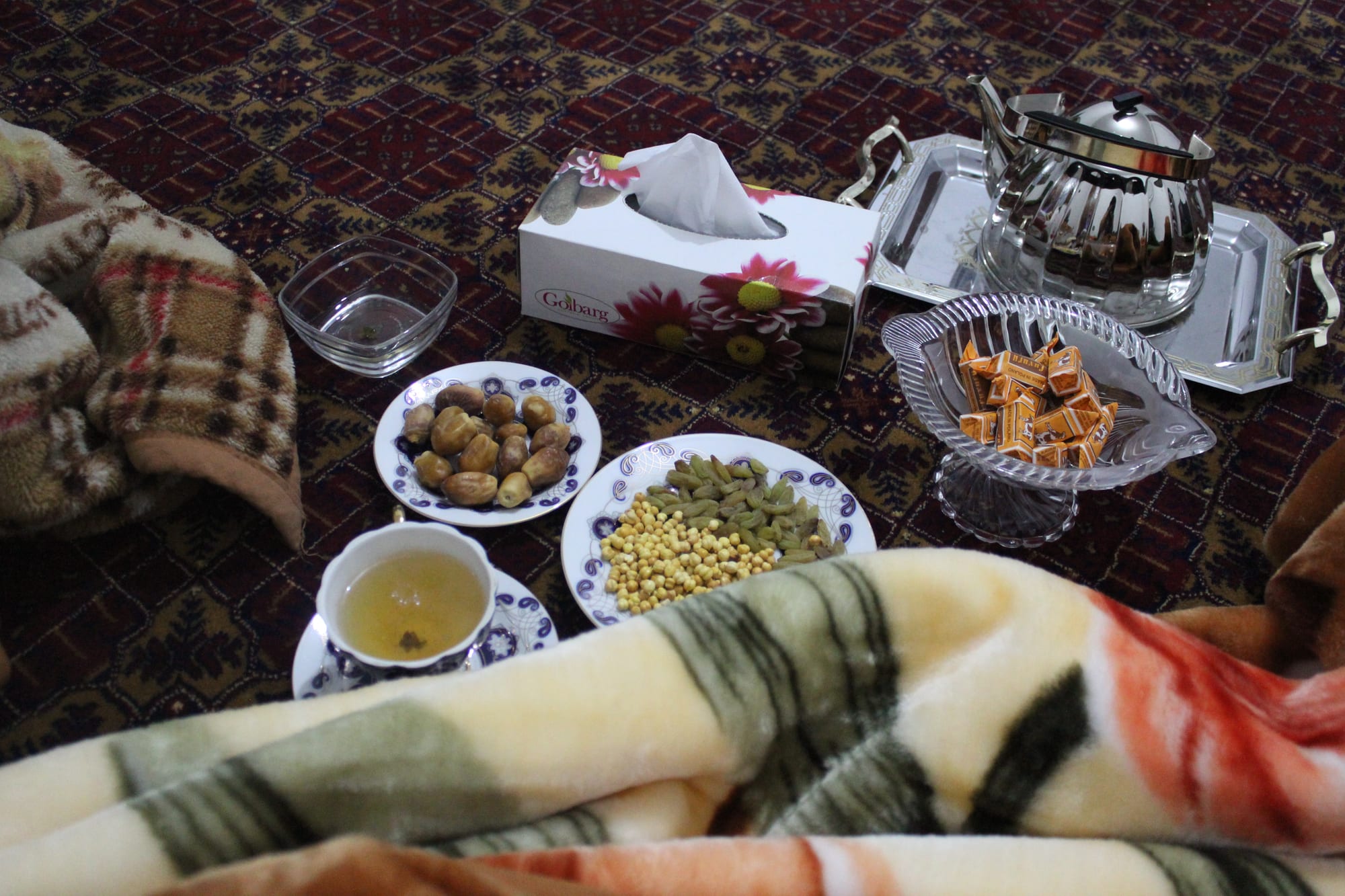 A metal teapot on a tray, a cup of green tea, and small plates of dried figs, cardamom pods and nuts set on a patterned cloth on the floor
