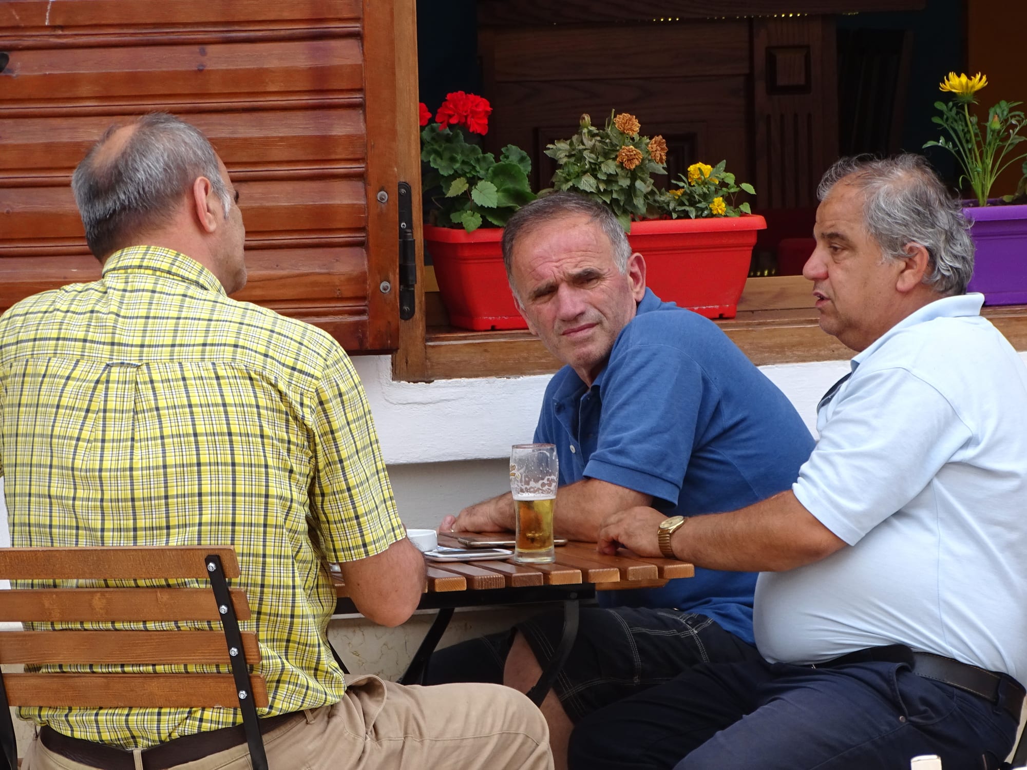 Three men sitting and talking at an outdoor café table with coffee and a beer