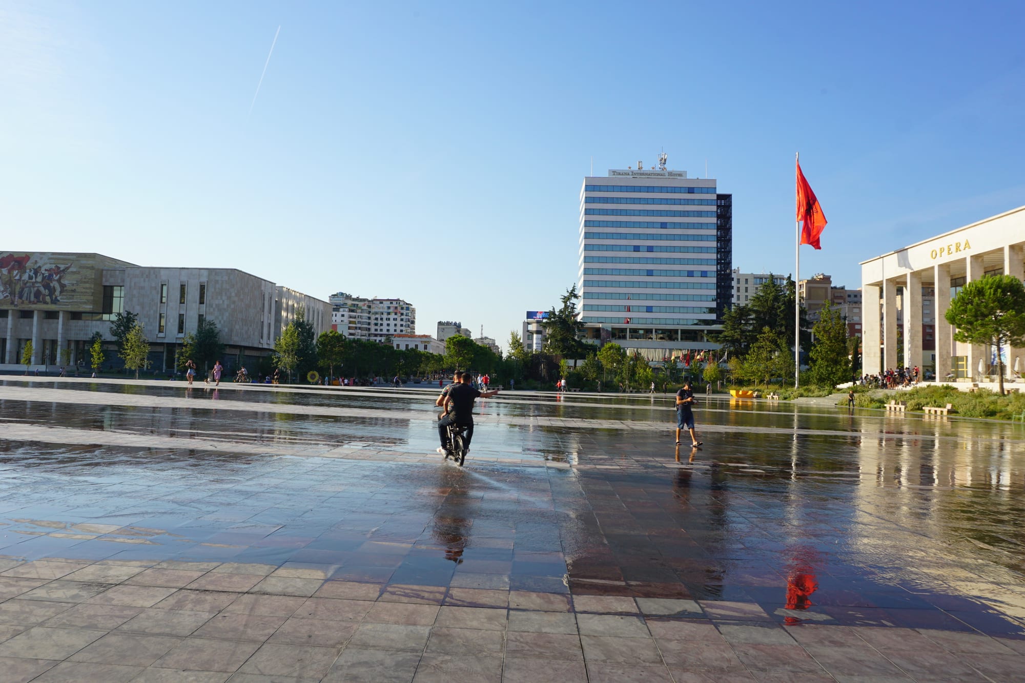 A vast open paved square with a mounted statue, fountains, and mountains rising behind the city