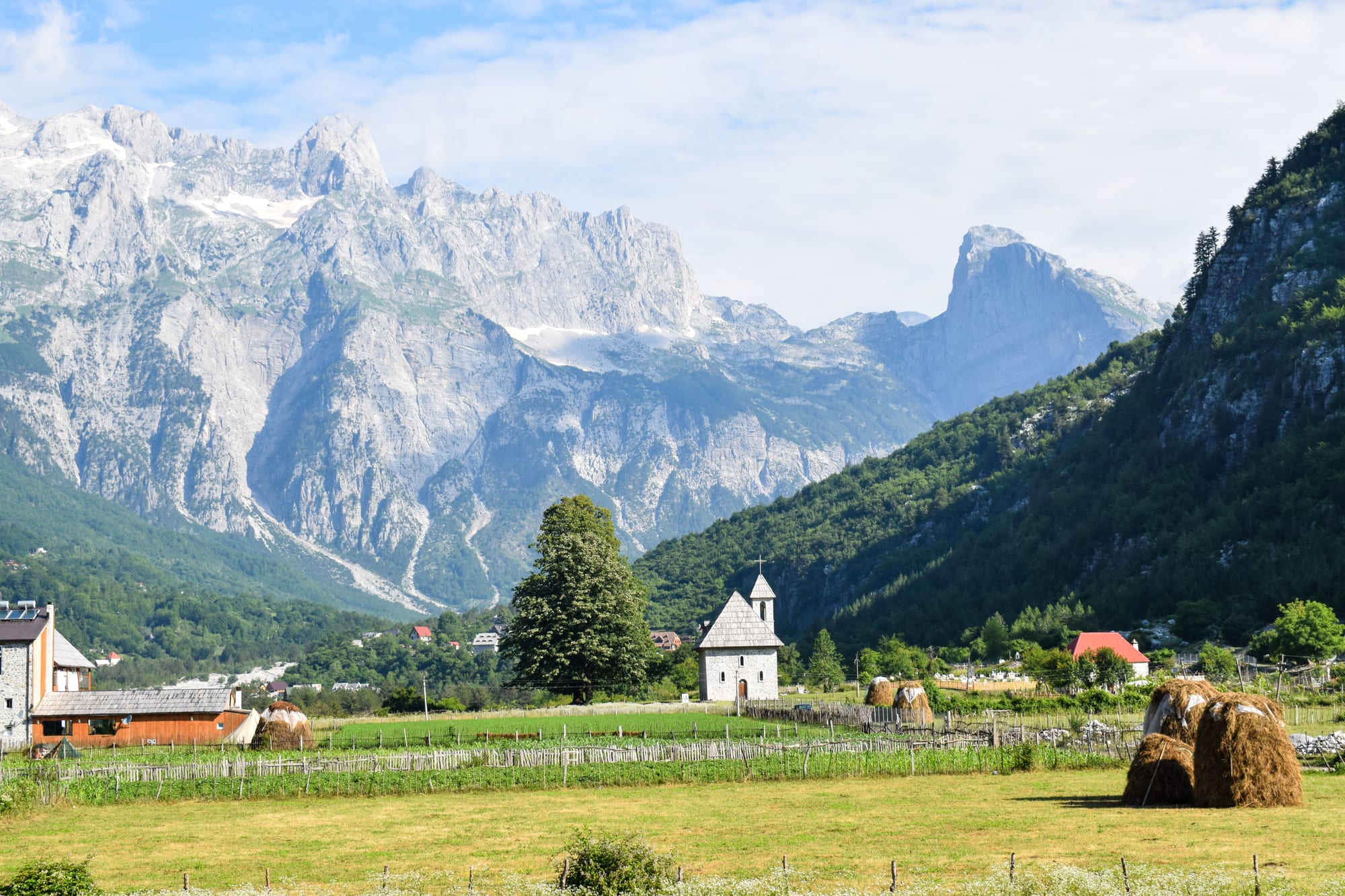 A small stone church with a shingled roof alone in a green valley beneath jagged grey peaks