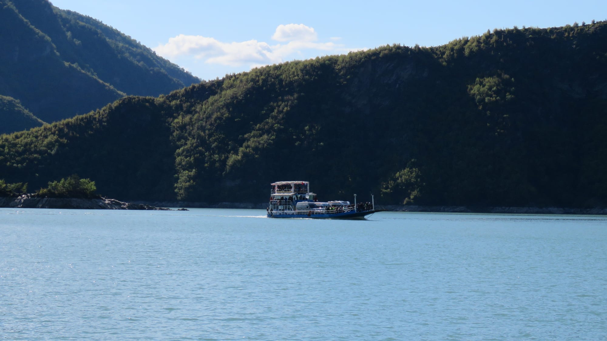 A small car ferry crossing a narrow green lake between very steep forested mountain walls
