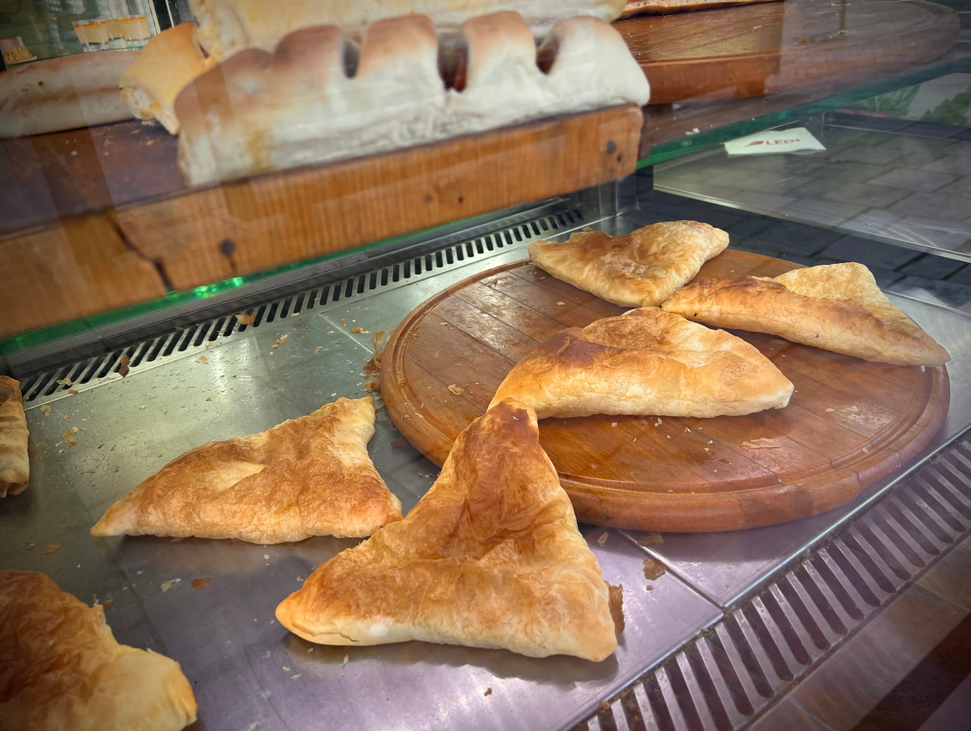 Golden triangular pastries on a round wooden board in a bakery display case, with a loaf of bread behind