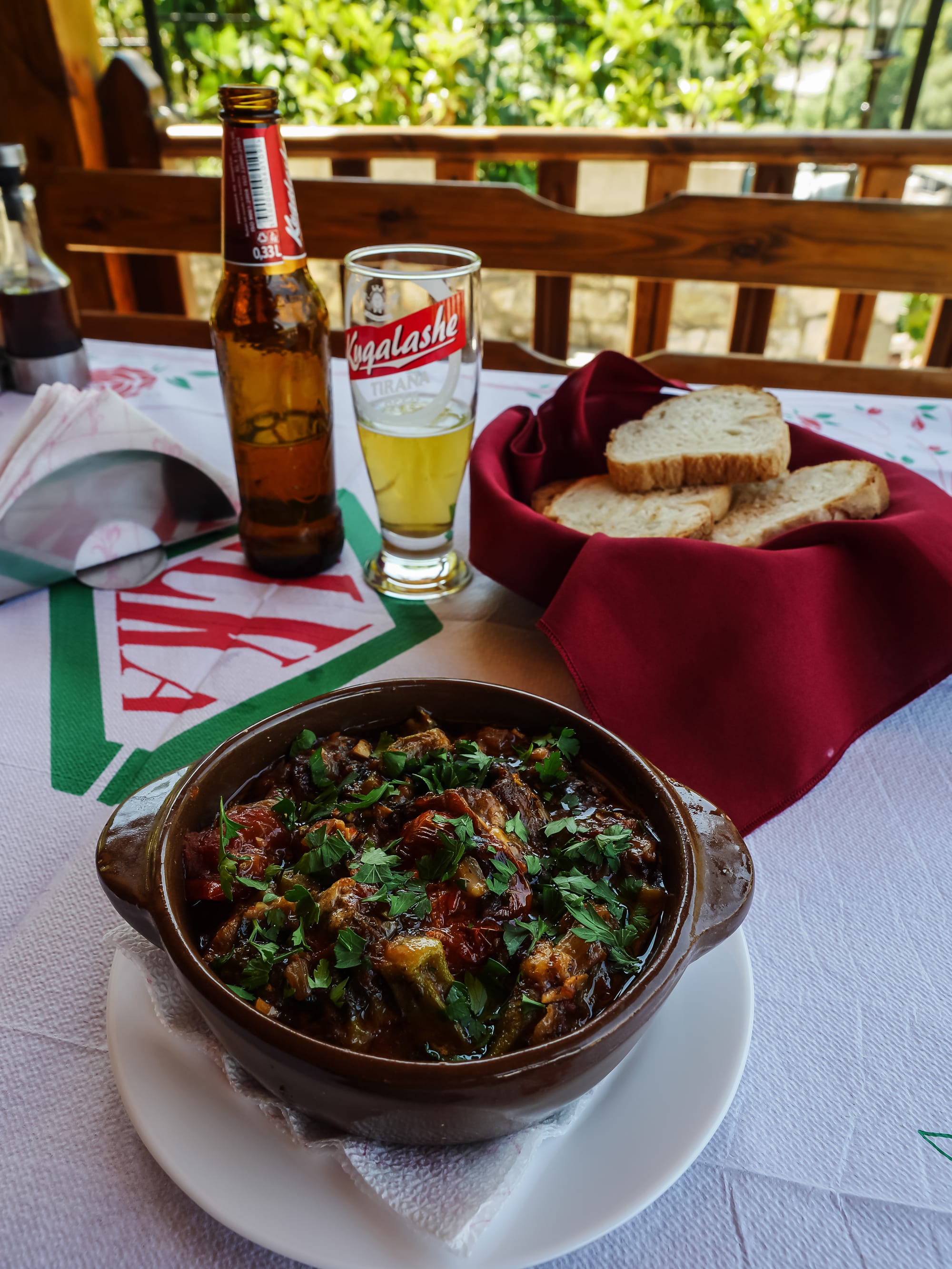 A brown clay pot of herb-topped pepper-and-tomato stew on a white plate, with a basket of sliced bread and a bottle and glass of pale beer, on a vine-shaded terrace