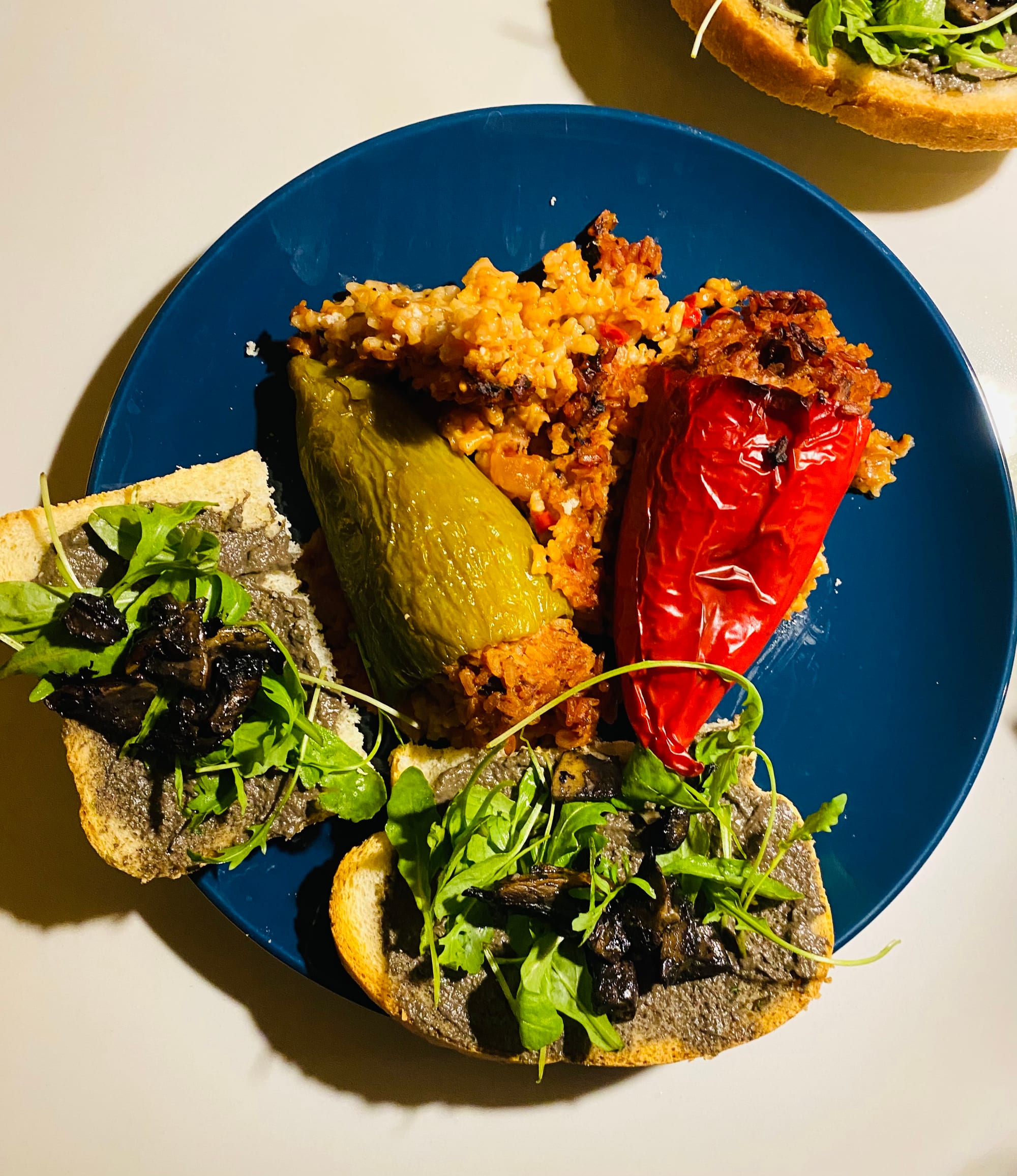 A blue plate with a green and a red pepper stuffed with seasoned rice, beside slices of bread topped with a dark spread and rocket
