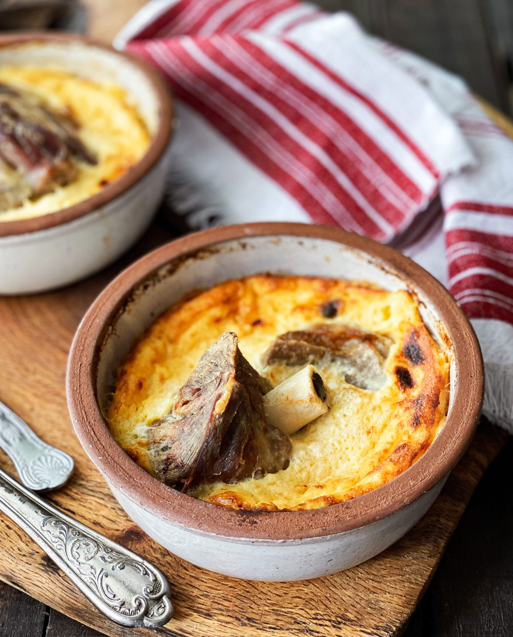 Two rustic ceramic bowls of baked yoghurt custard, golden and puffed on top, with pieces of lamb on the bone showing through