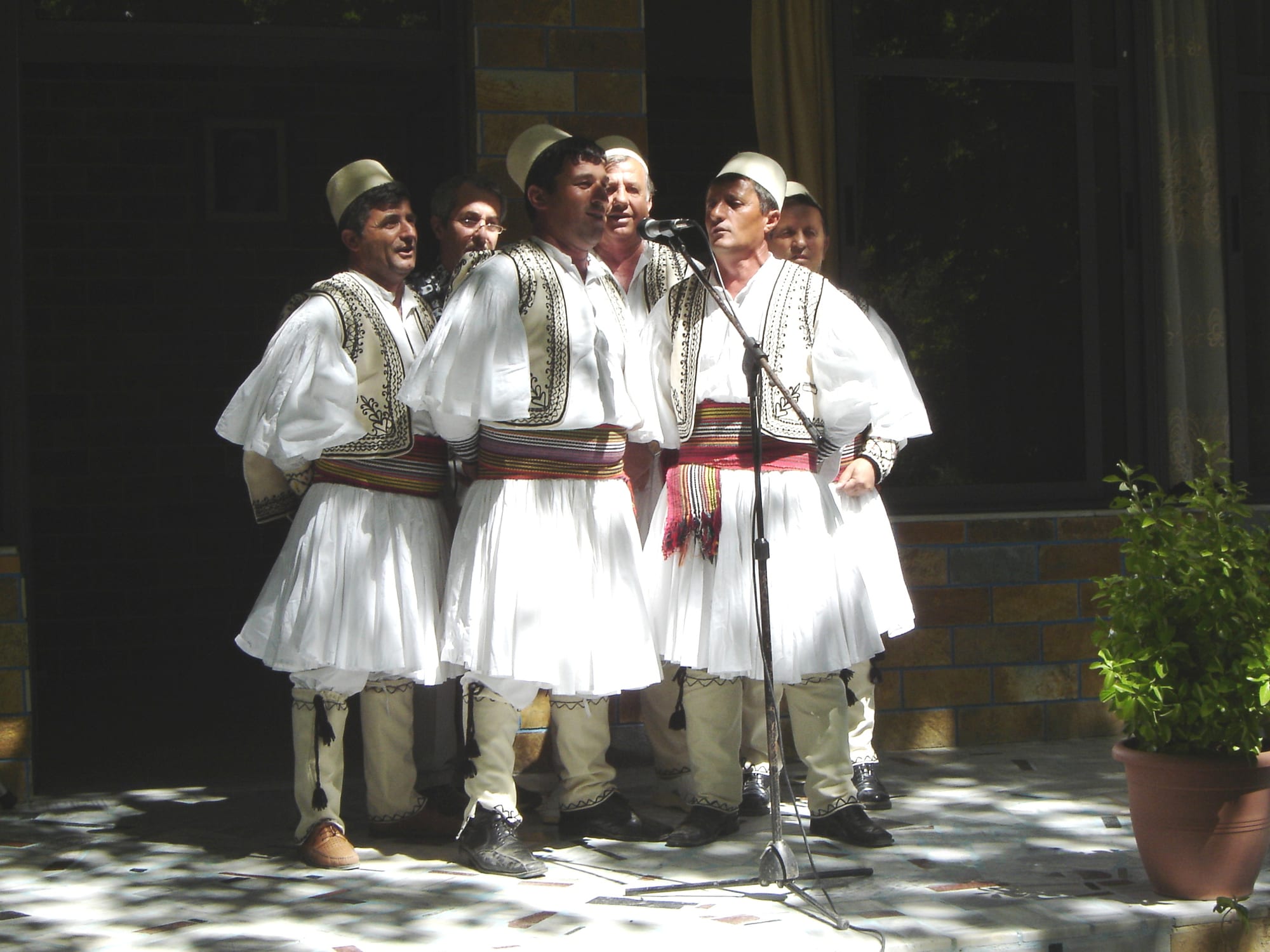 A group of men in traditional dress, with pleated white kilts, embroidered black waistcoats, red sashes and white felt caps, singing together around a microphone on a sunlit terrace