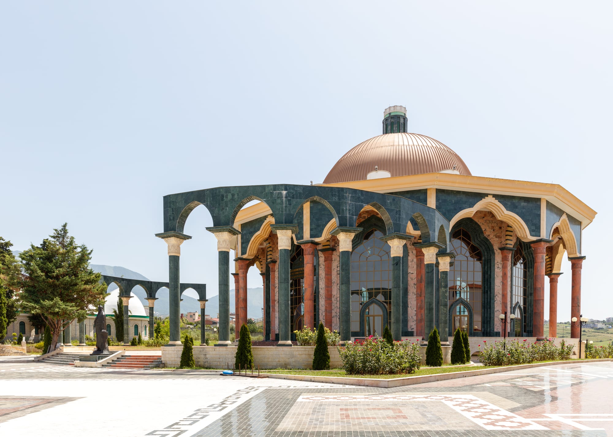 An octagonal building with a copper dome, green marble arches and red columns, set in a paved courtyard with a statue and more arcades behind