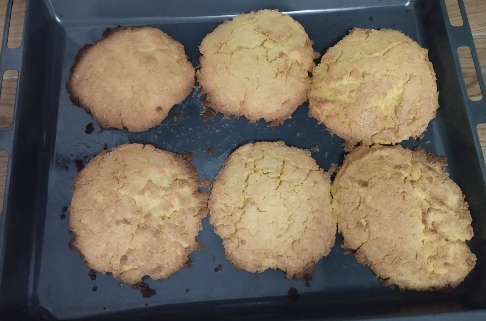 Six round, golden biscuits with cracked tops and browned edges on a dark baking tray