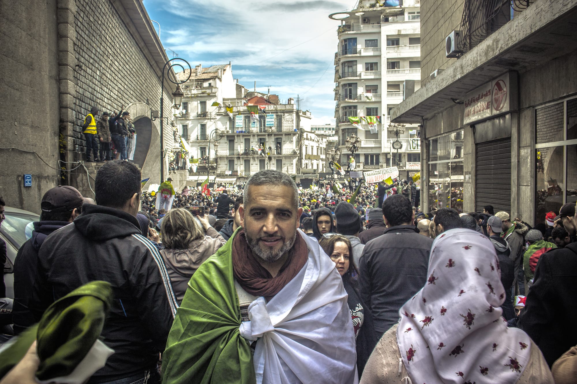 A dense crowd filling a wide city avenue, many people waving Algerian flags