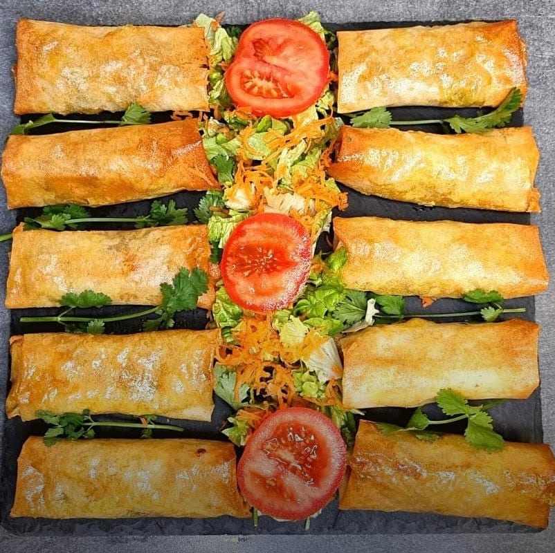 Ten golden fried pastry rolls arranged in two rows around a strip of shredded lettuce, carrot and tomato slices on a dark slate board