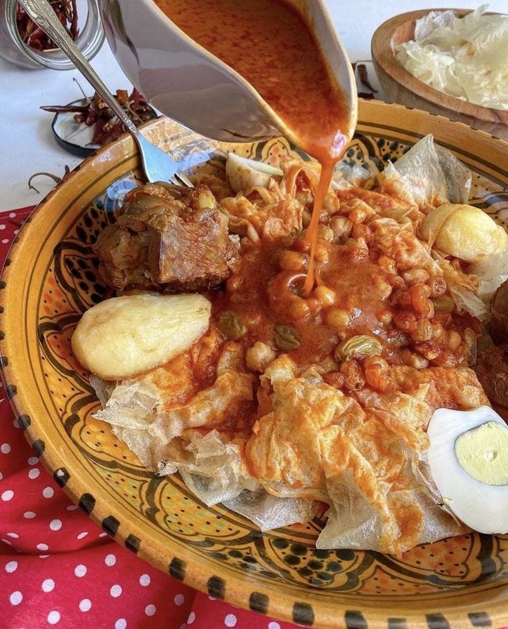 A patterned bowl of torn flatbread pieces with a rich orange-red sauce being poured over, chickpeas, raisins, a boiled egg and pieces of meat visible
