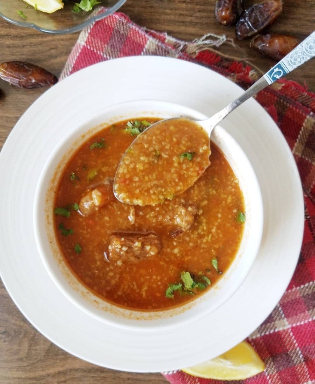 An overhead view of a bowl of orange, thick soup with a spoonful lifted showing cracked wheat and pieces of meat, dates and lemon wedges on the tablecloth beside it