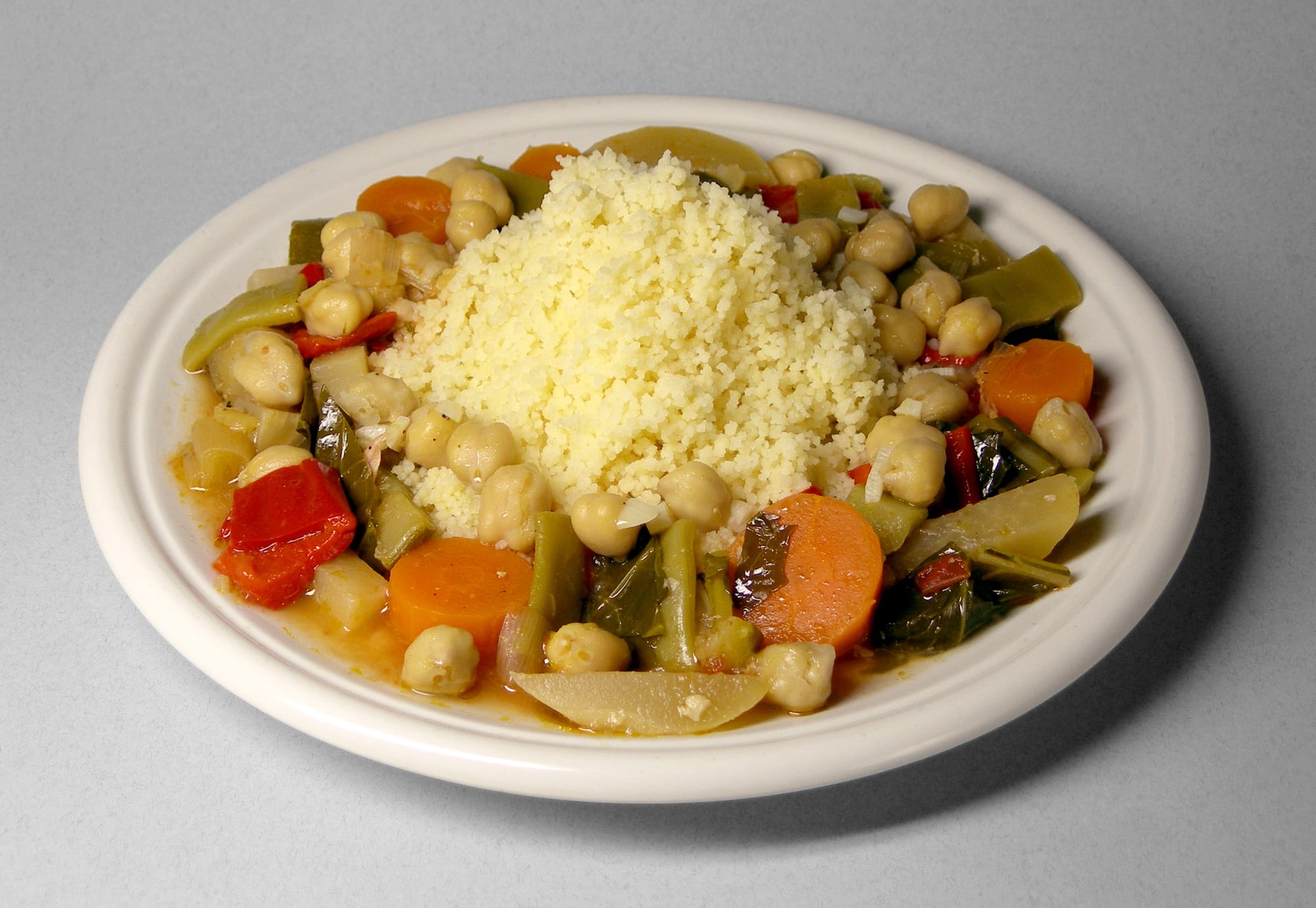 A white plate of pale steamed couscous surrounded by a stew of carrots, chickpeas, green beans and peppers in broth