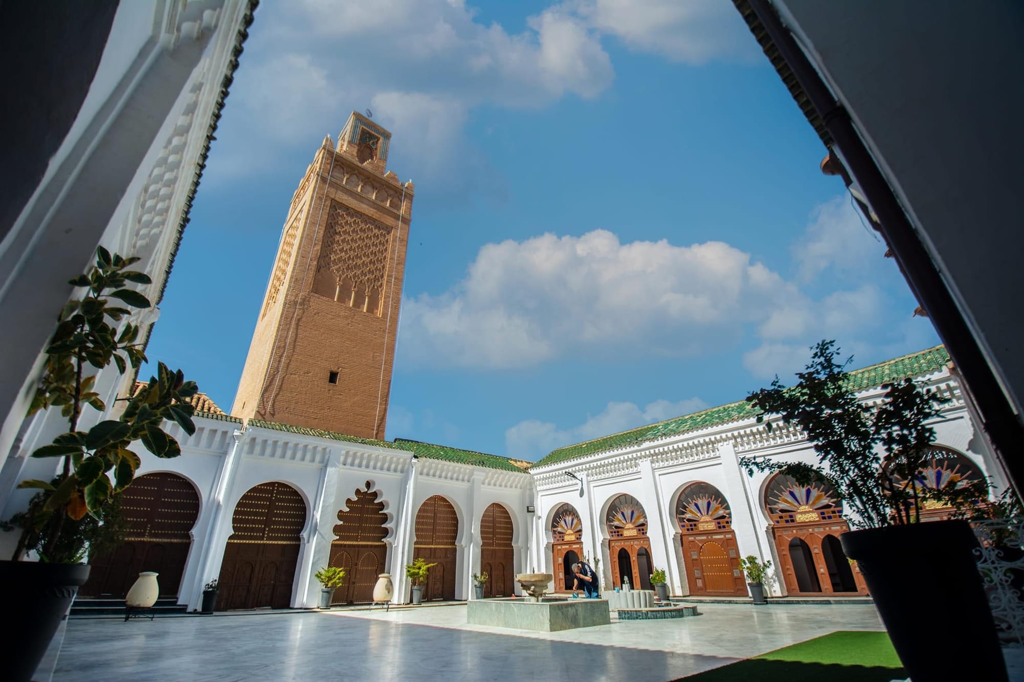 A whitewashed courtyard with carved wooden doors under horseshoe arches, a green-tiled roofline and a tall tan brick minaret against a blue sky