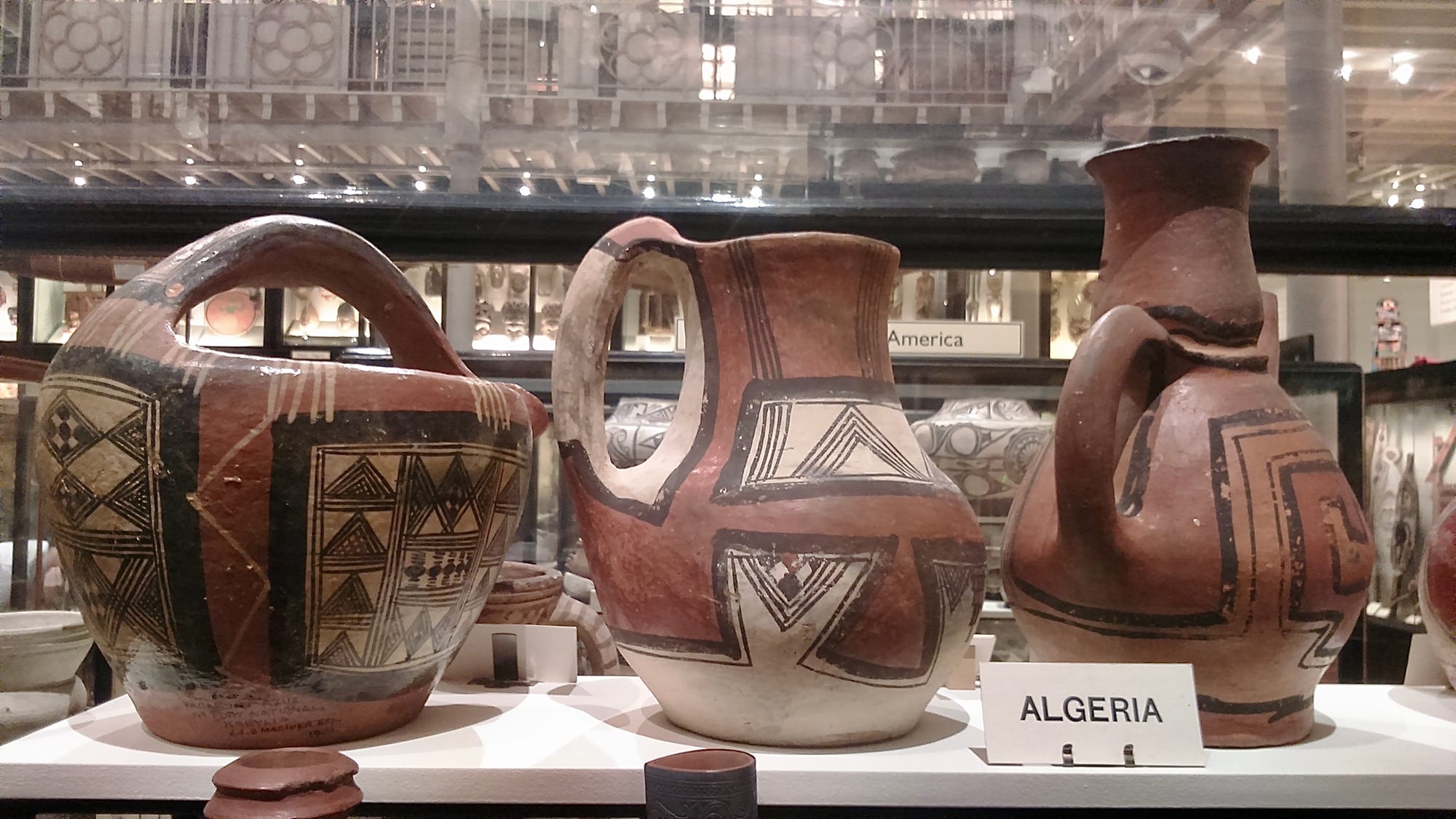 Three handmade earthenware vessels, a large jar with a handle, a jug and a tall pitcher, painted in red and cream with black geometric triangles and lines, in a museum case labelled Algeria