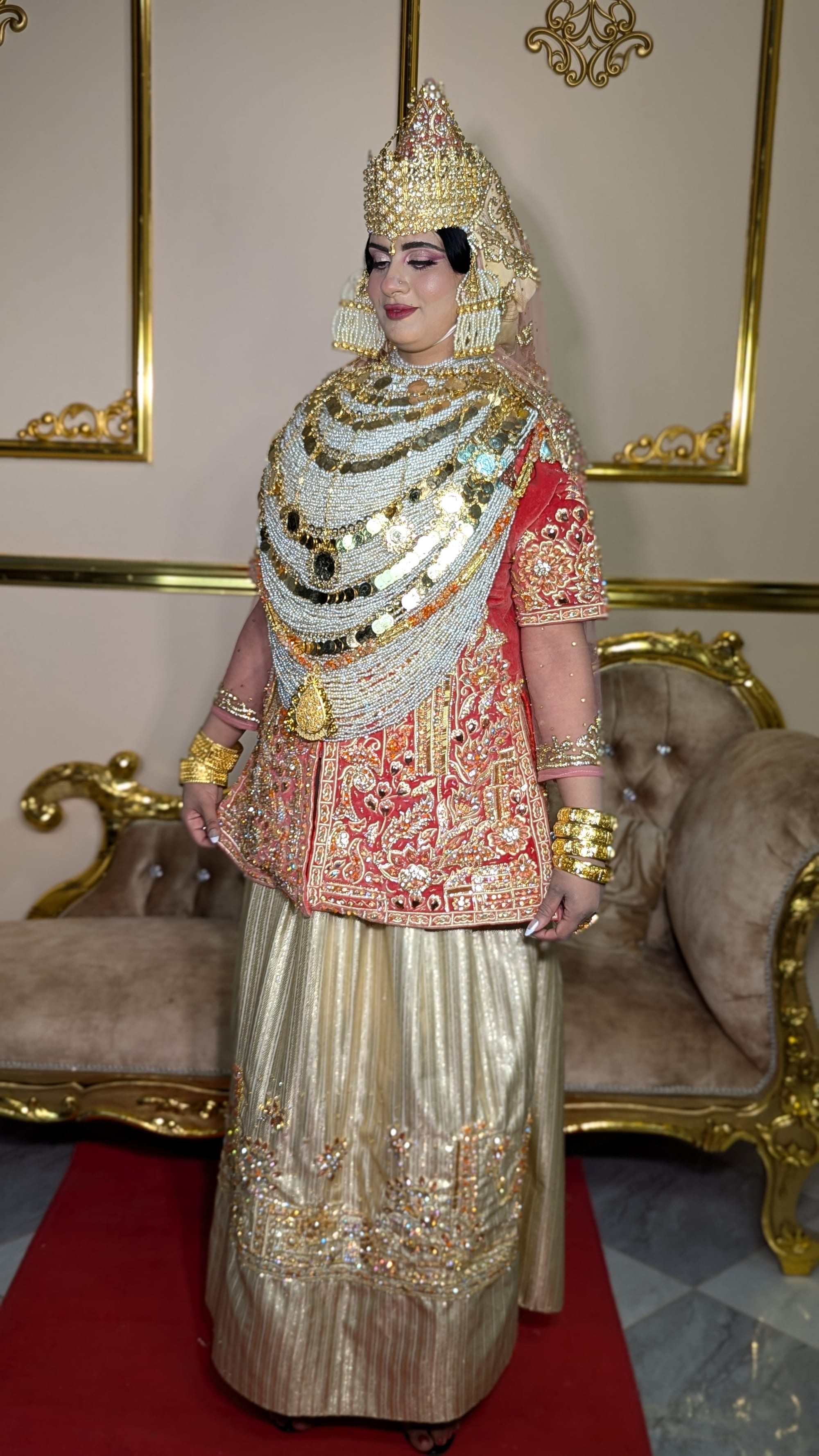 A bride in a tall gold crown-like headdress, strands of pearls and gold coins draped over a red velvet caftan embroidered in gold, and a long pleated gold skirt, standing in front of a gilded sofa