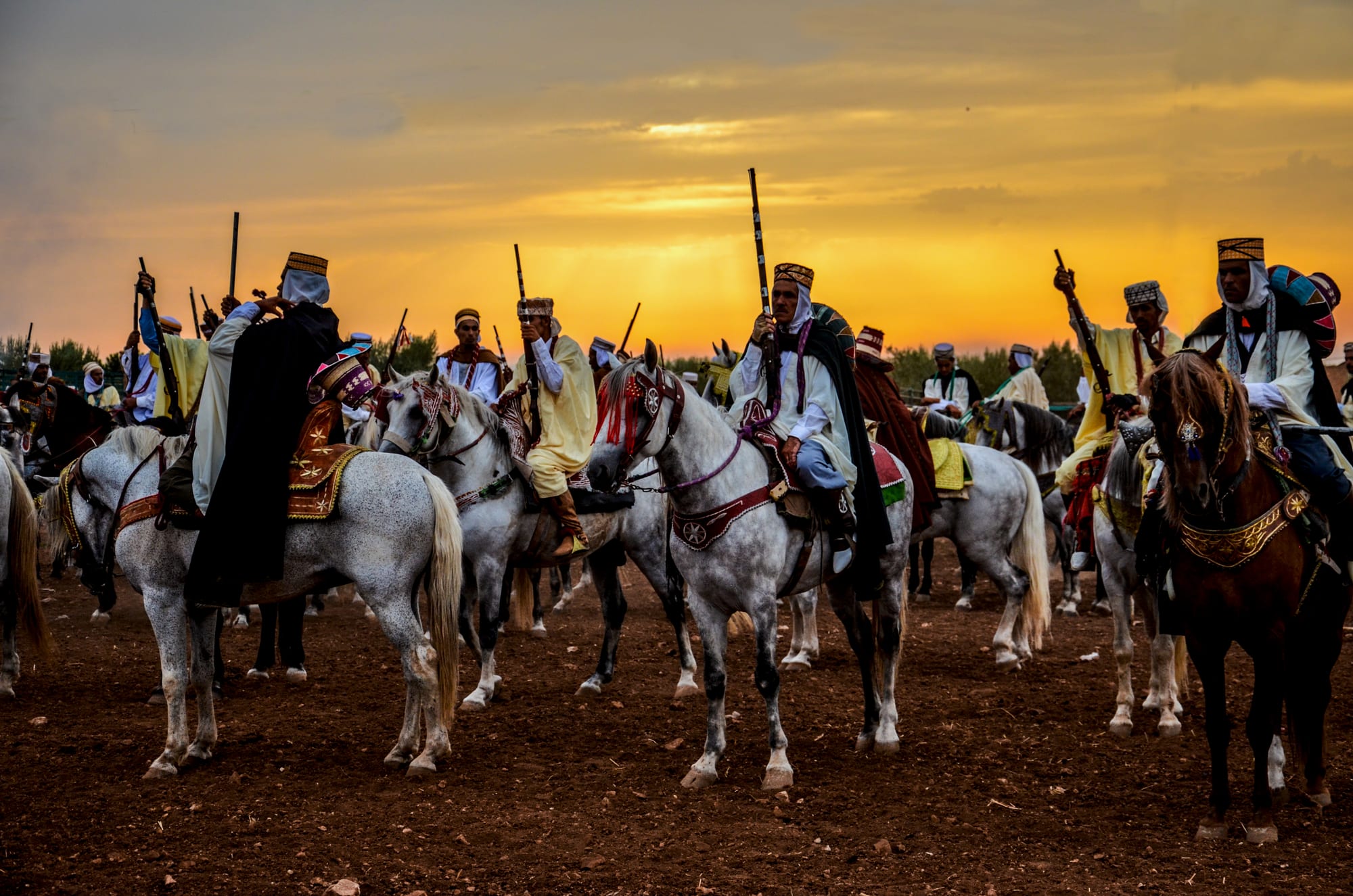 A line of horsemen in white robes, embroidered caps and dark cloaks sit on decorated grey horses holding long rifles upright against an orange sunset sky