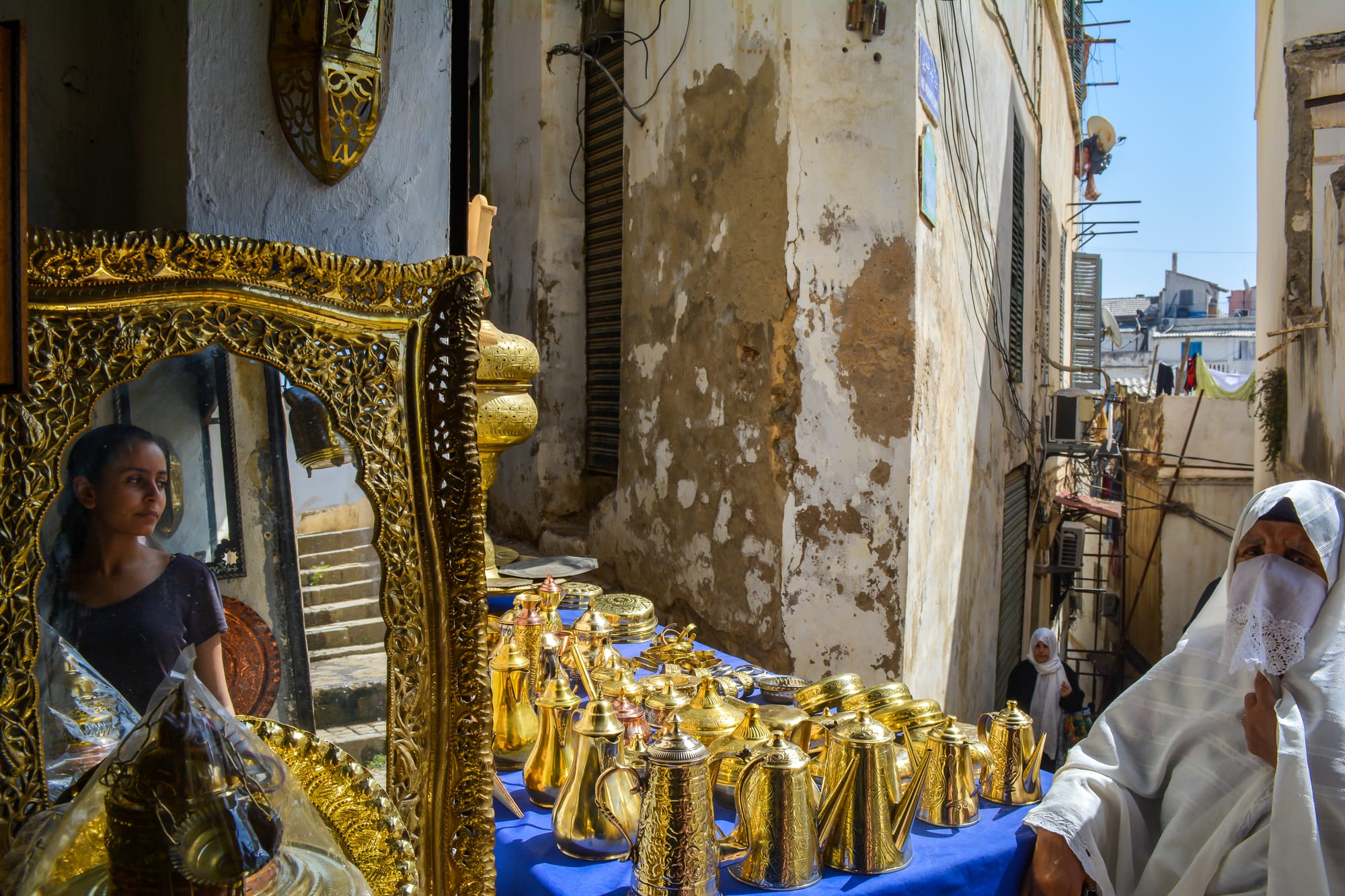 A narrow alley in the Casbah of Algiers with a stall of brass and metalware and two women in white headscarves passing by