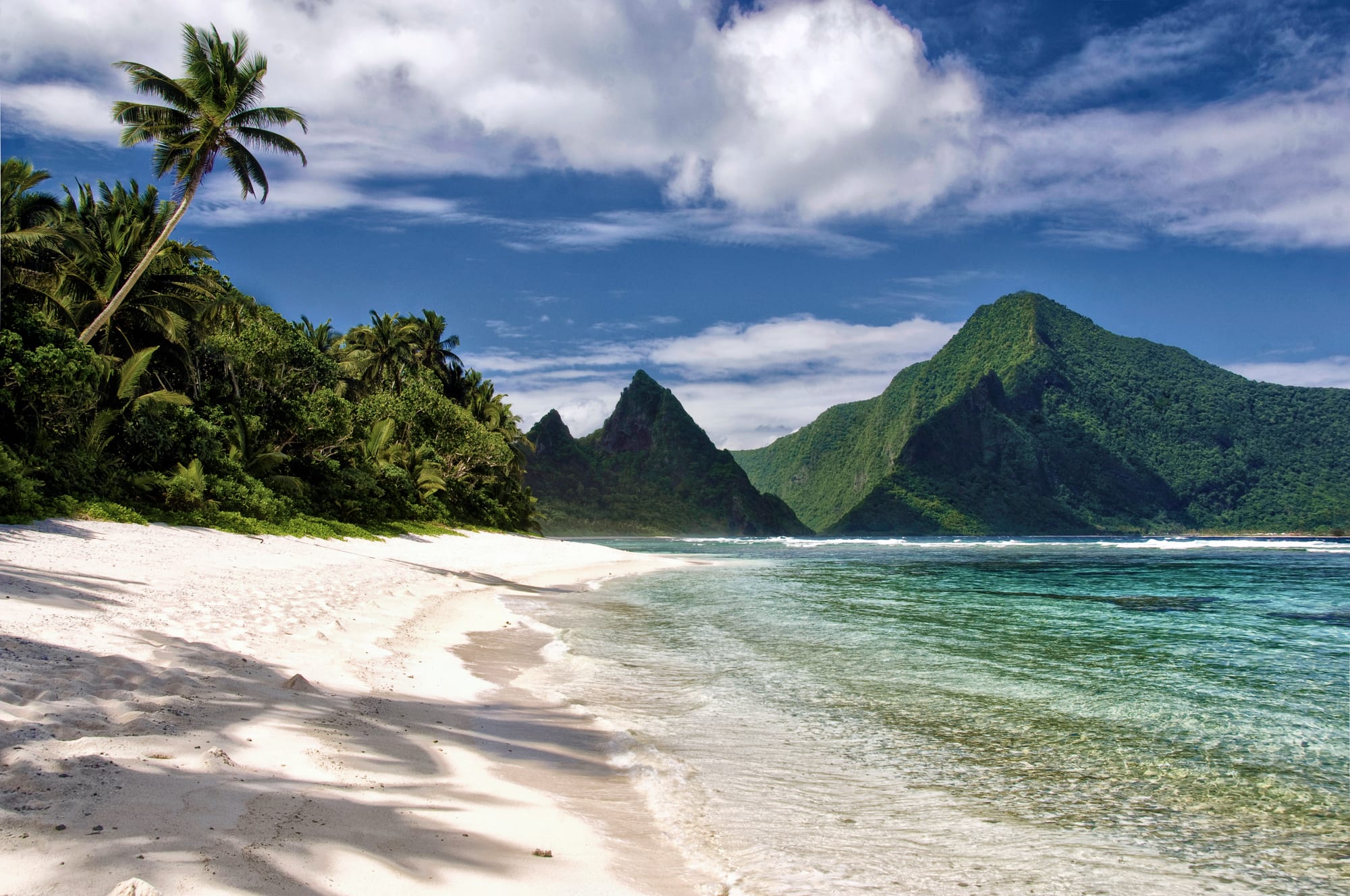 A curving white beach backed by jungle-covered peaks, reef visible offshore