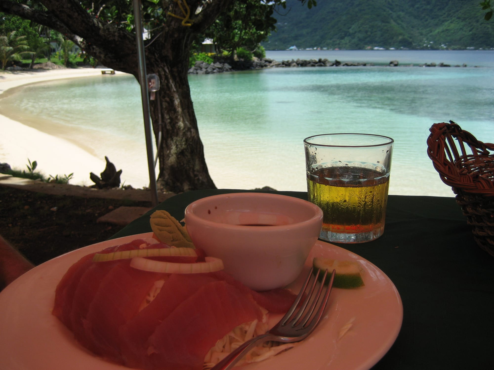 A plate of sliced raw red tuna with a small bowl of dipping sauce and a glass of beer on a table under a tree, with a pale turquoise bay, a sandy beach and green hills behind