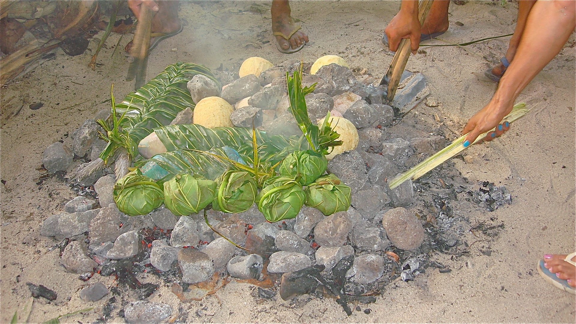 Small leaf-wrapped parcels and pale round pieces of breadfruit arranged on a bed of hot volcanic stones on a sandy beach, with people tending the fire