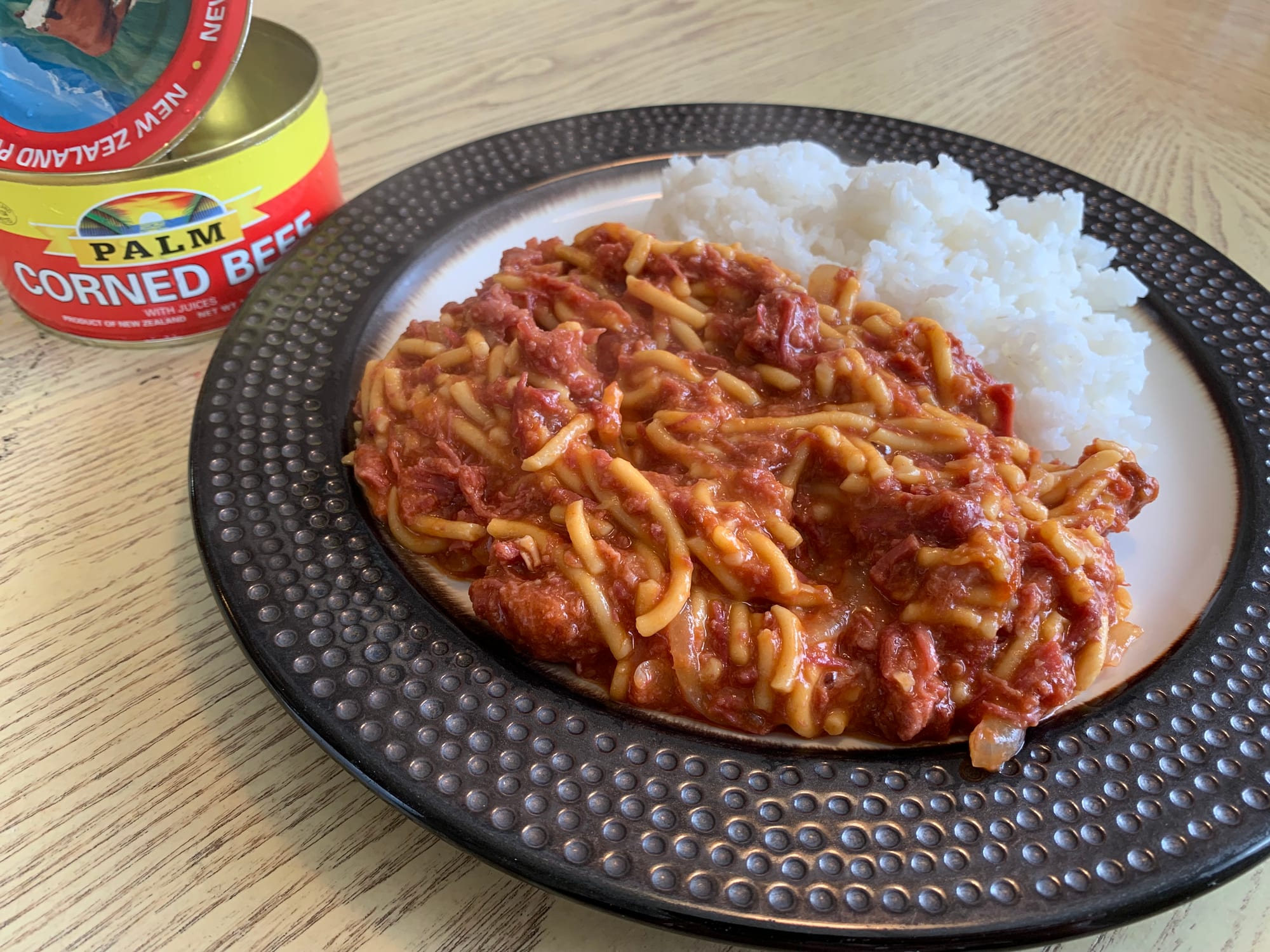 A plate of pasta in red sauce mixed with meat, served with white rice, and a labelled tin of corned beef on the table beside it