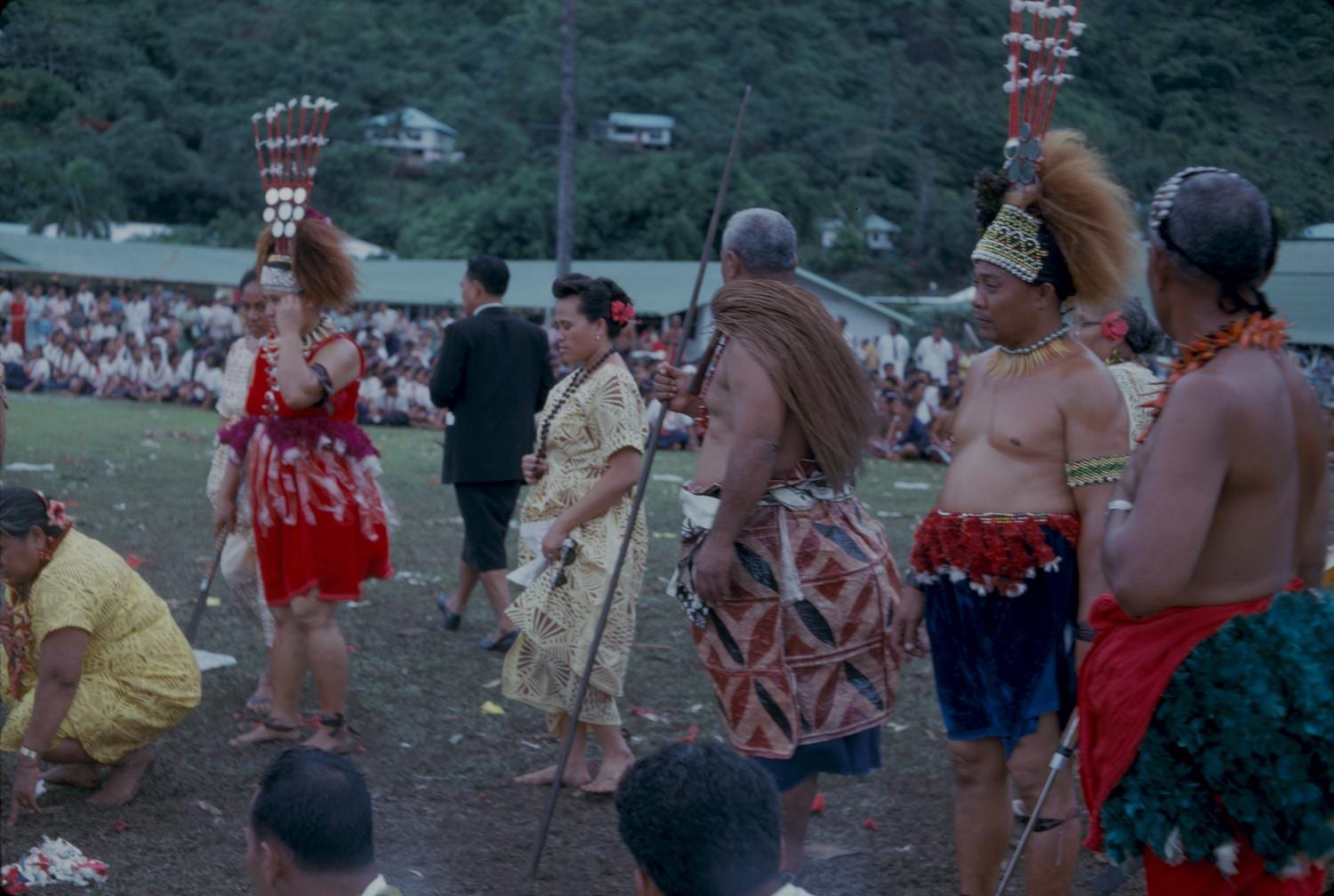 Participants in traditional dress on a grassy field, including a woman in a red costume and tall feathered headdress, a man wearing a patterned wrap and a long fibre garment, and another man in a tall headdress, with a crowd behind