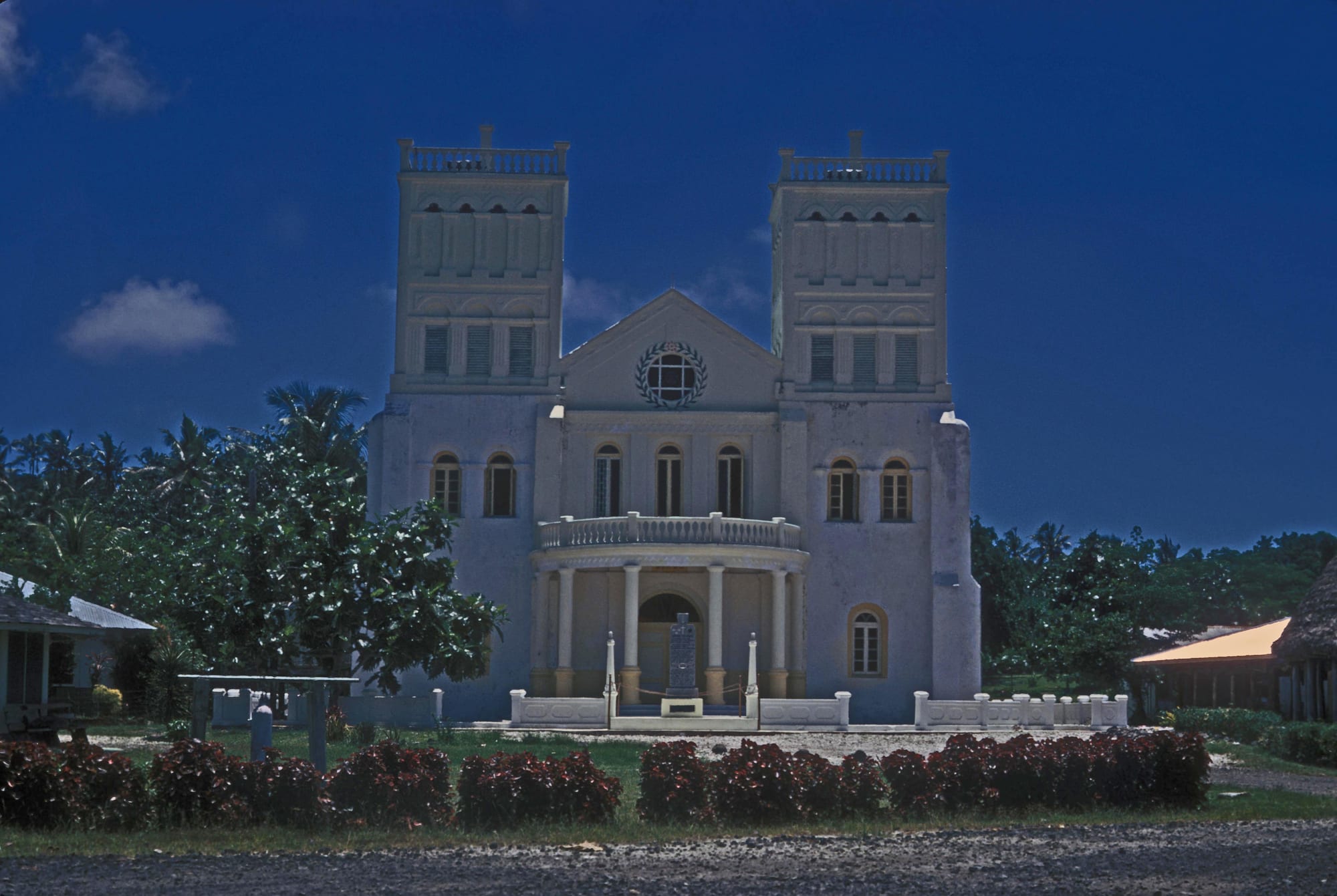 A large pale church with two square towers, a columned porch and a stone memorial in front, set among tropical trees under a deep blue sky
