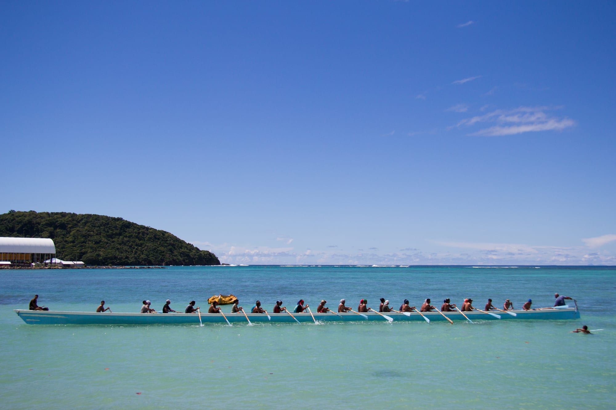 A long pale blue rowing boat with a row of more than twenty rowers pulling oars across a turquoise lagoon under a clear blue sky, with a wooded headland behind