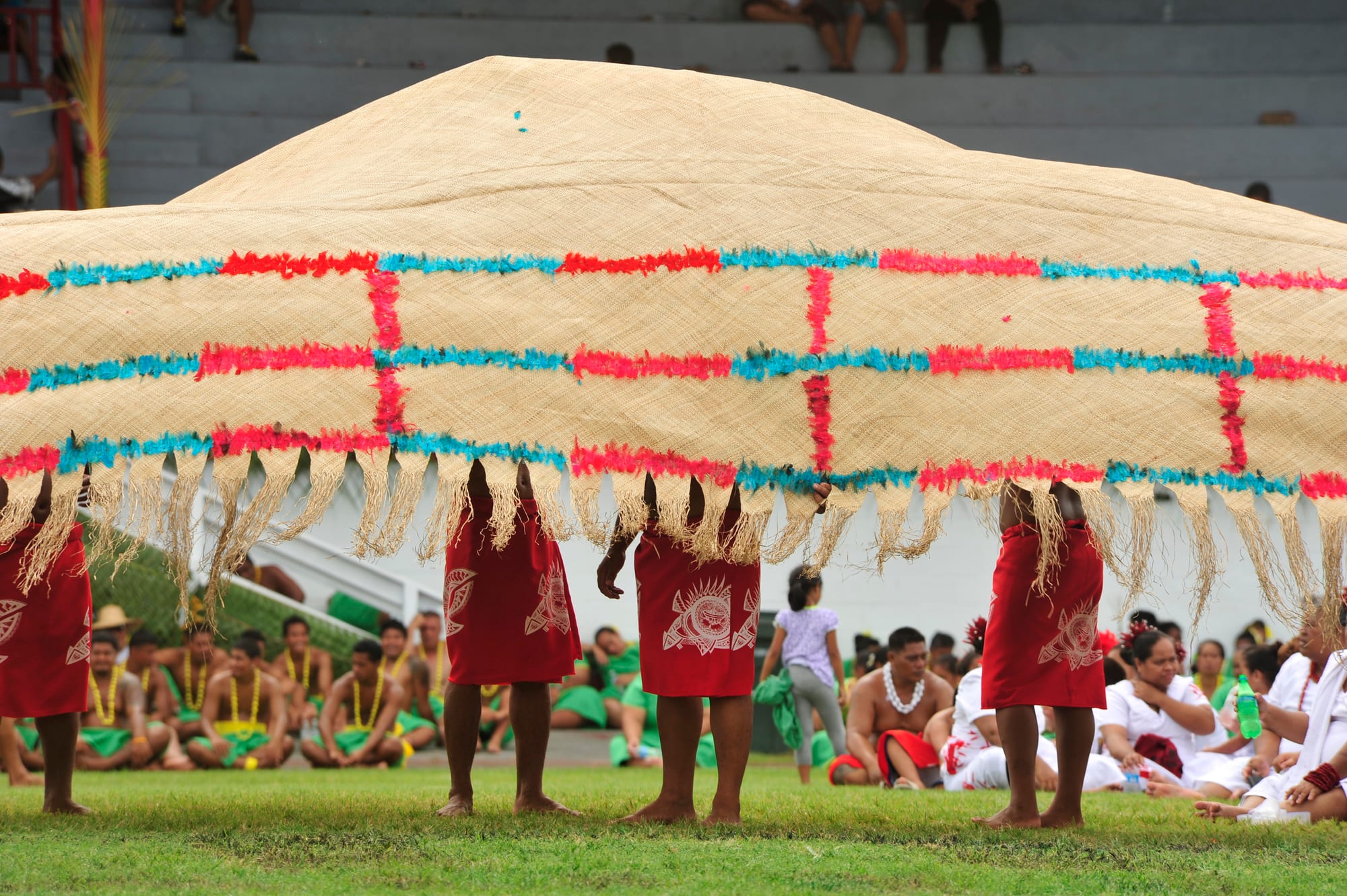 A huge pale woven mat edged with rows of red and turquoise feathers is held up high, hiding the faces of the people beneath it, who wear red wraps, with a seated crowd behind