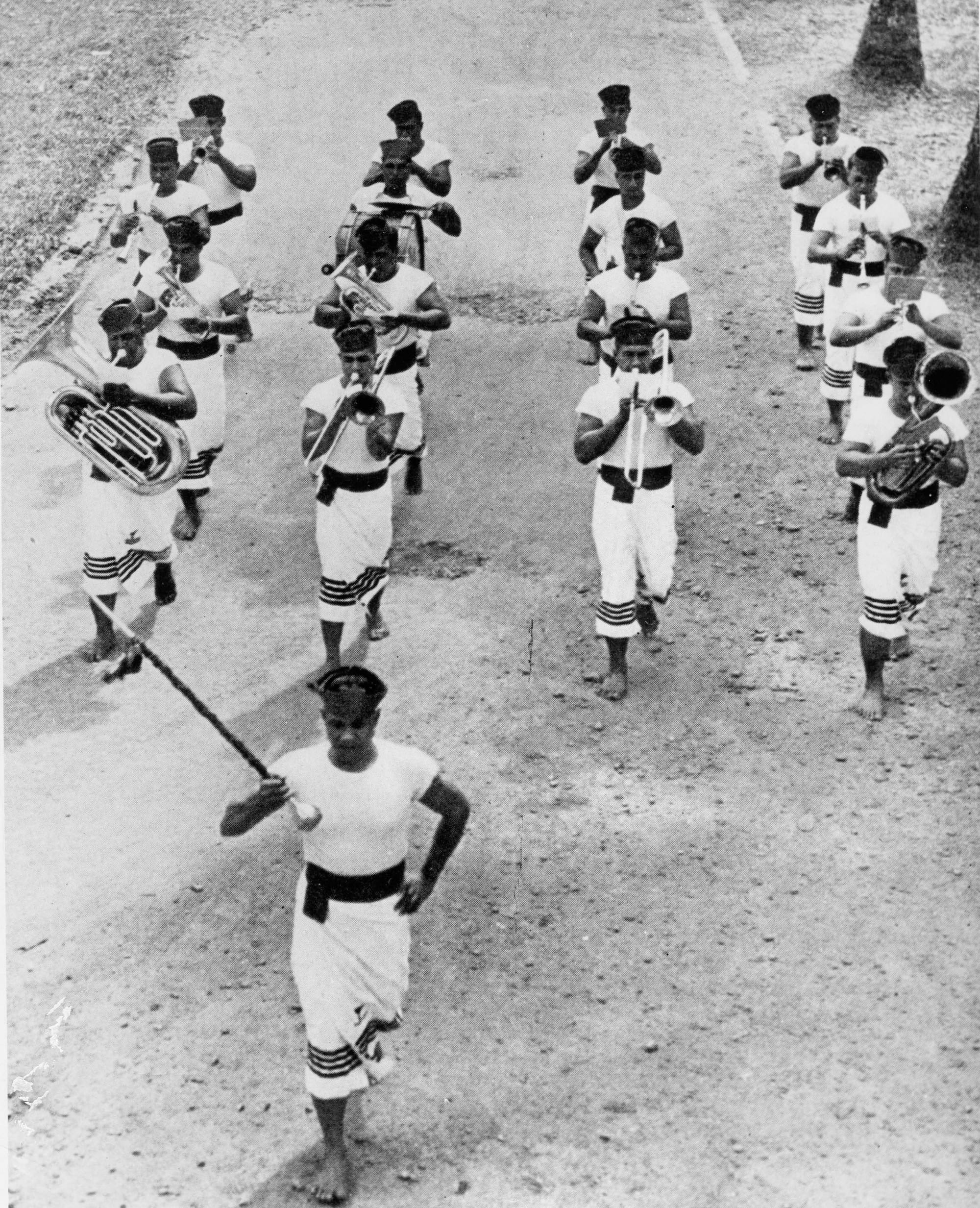 A black-and-white photograph, taken from above, of a barefoot brass band in white uniforms marching along a road behind a drum major