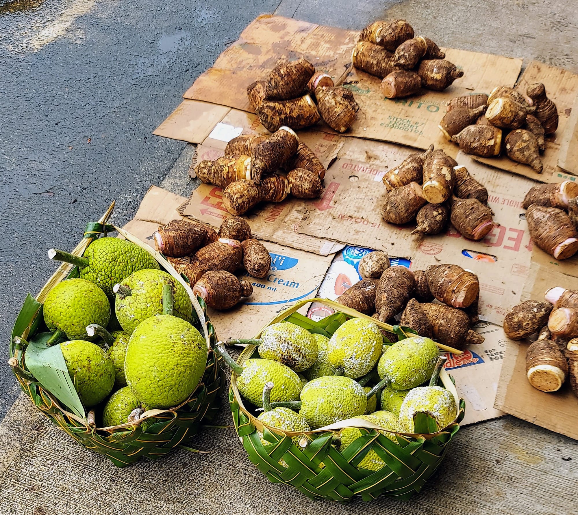 Woven baskets of breadfruit and piles of taro root laid out for sale on cardboard at the roadside