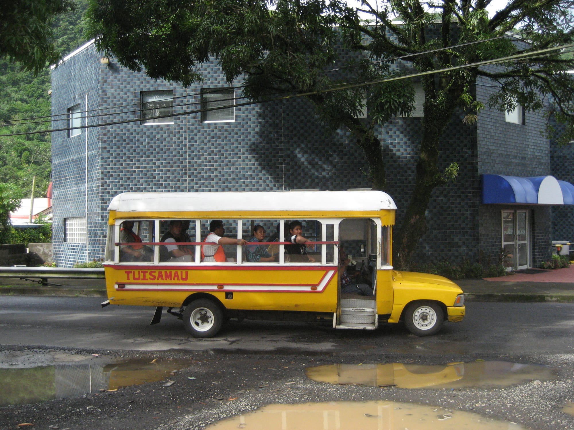 A bright yellow, hand-built wooden bus named Tuisamau carrying passengers along a street in Pago Pago