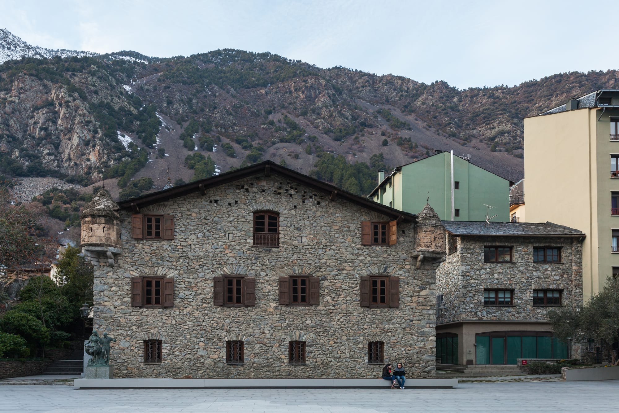 A small fortified stone house with a square tower and stepped gable, mountains behind