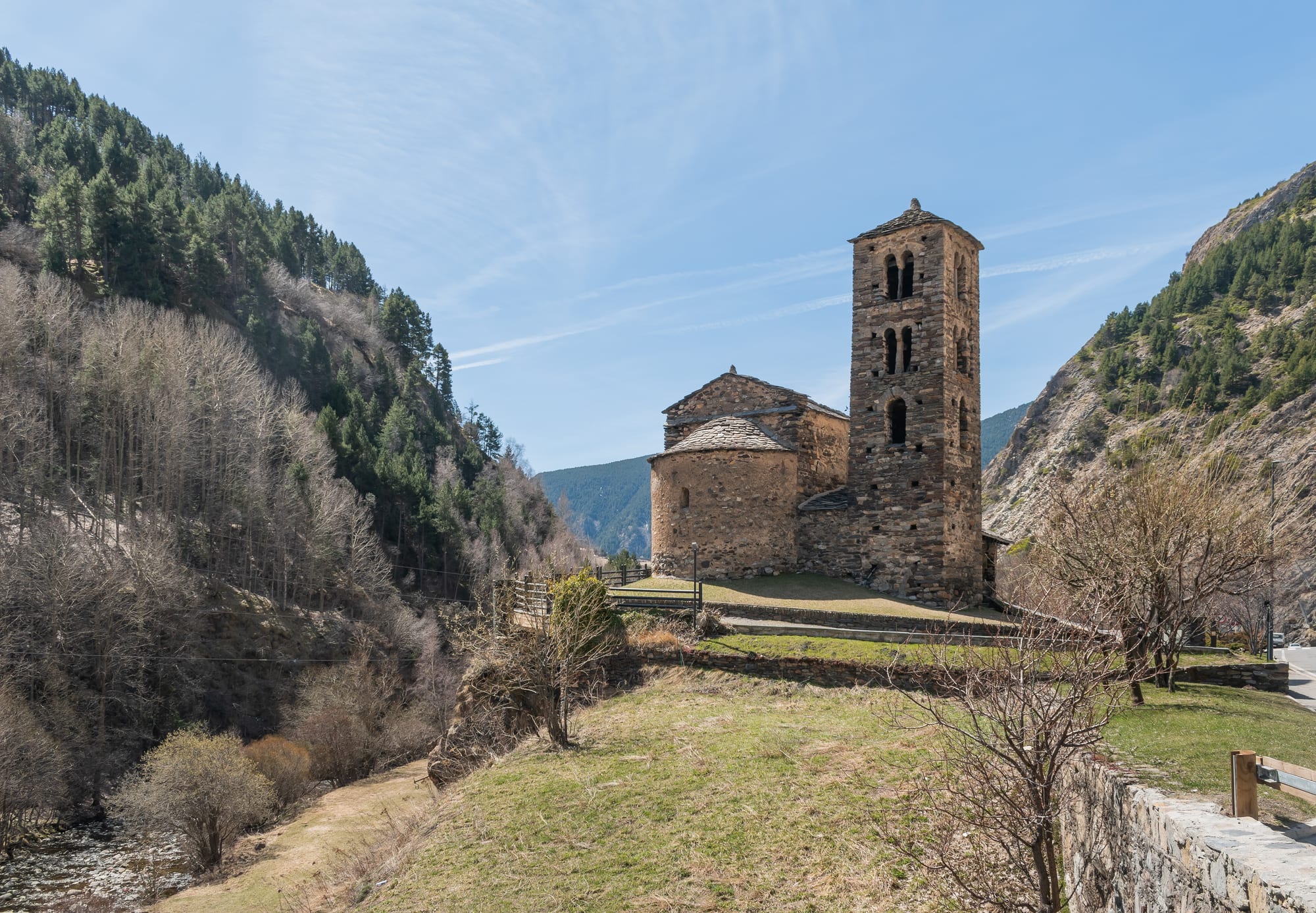 A stone church with a square tower and covered porch on a green terrace, snowy peaks behind