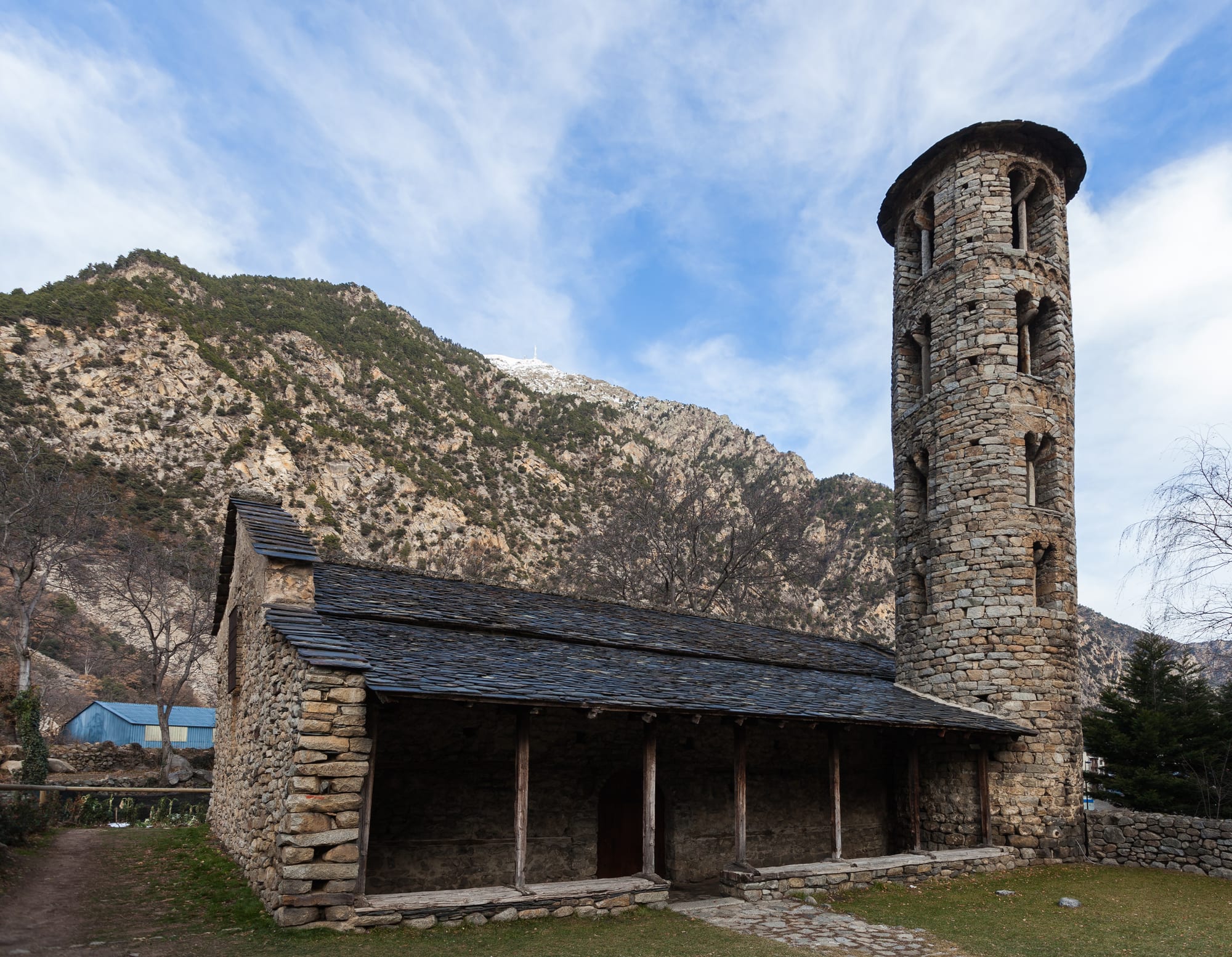 A small stone church with a tall round bell tower in a green valley below mountains