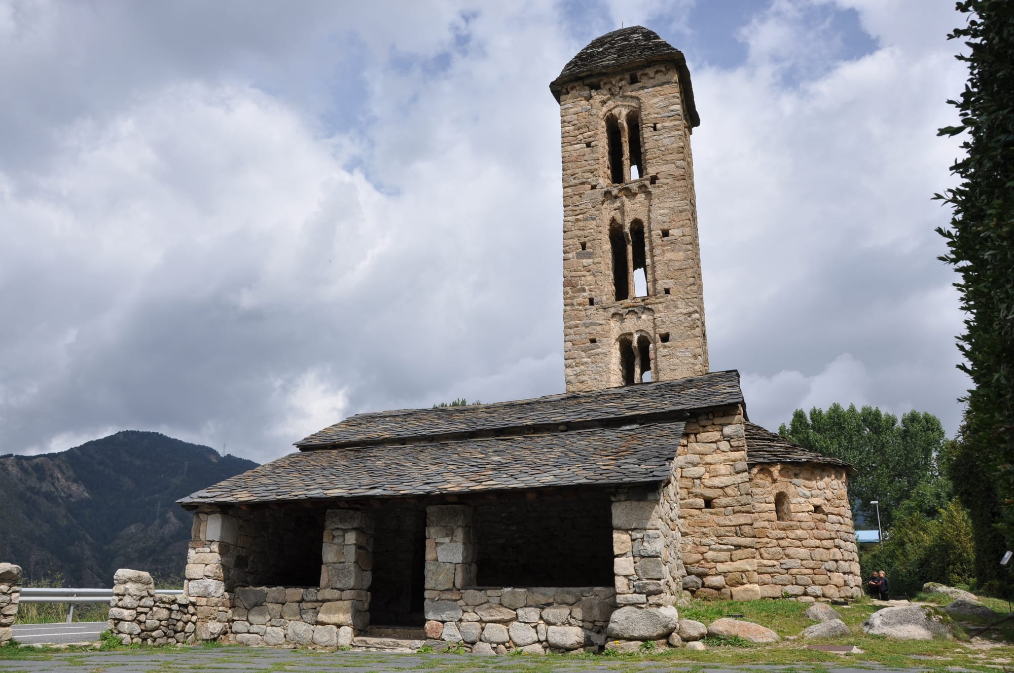 A small Romanesque church of rough stone with a slate roof, an open arcaded porch and a tall narrow bell tower pierced by paired arched windows, under a cloudy sky with a wooded mountain behind