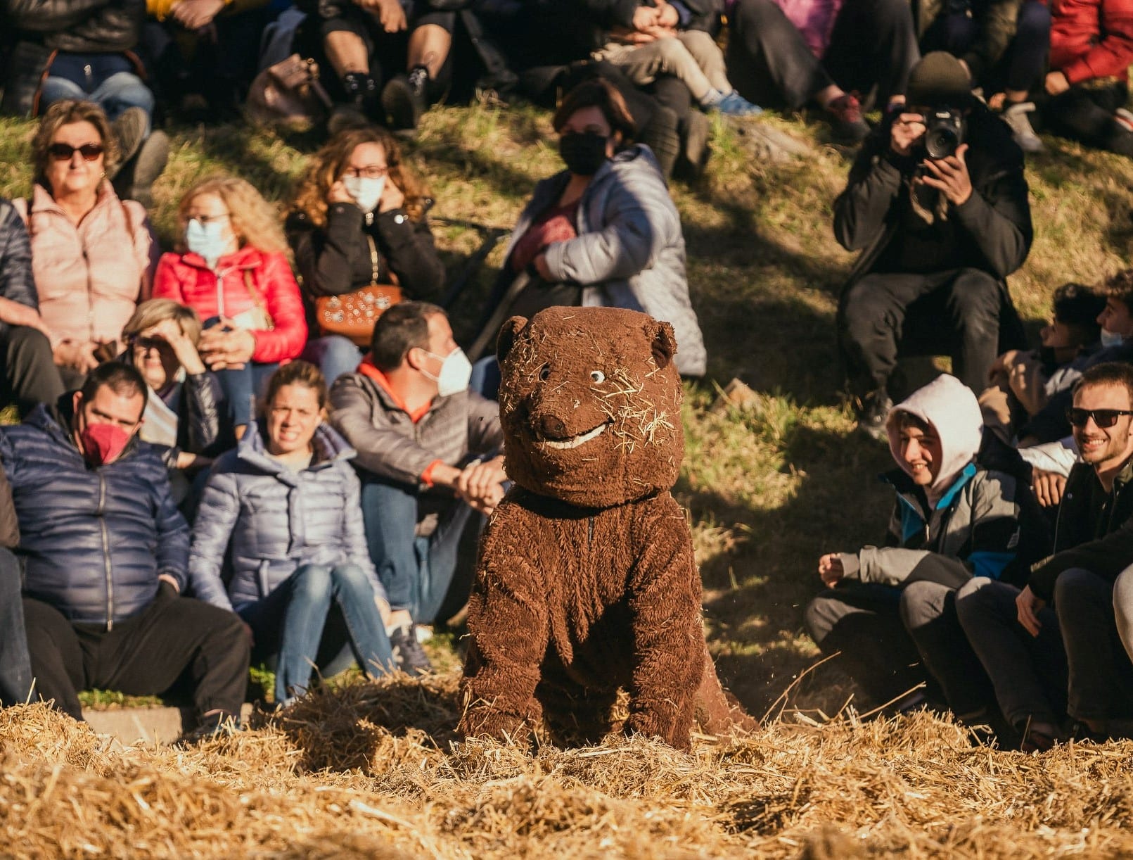 A performer in a shaggy brown bear costume crouches on a bed of straw in front of a hillside crowd of seated spectators, several smiling