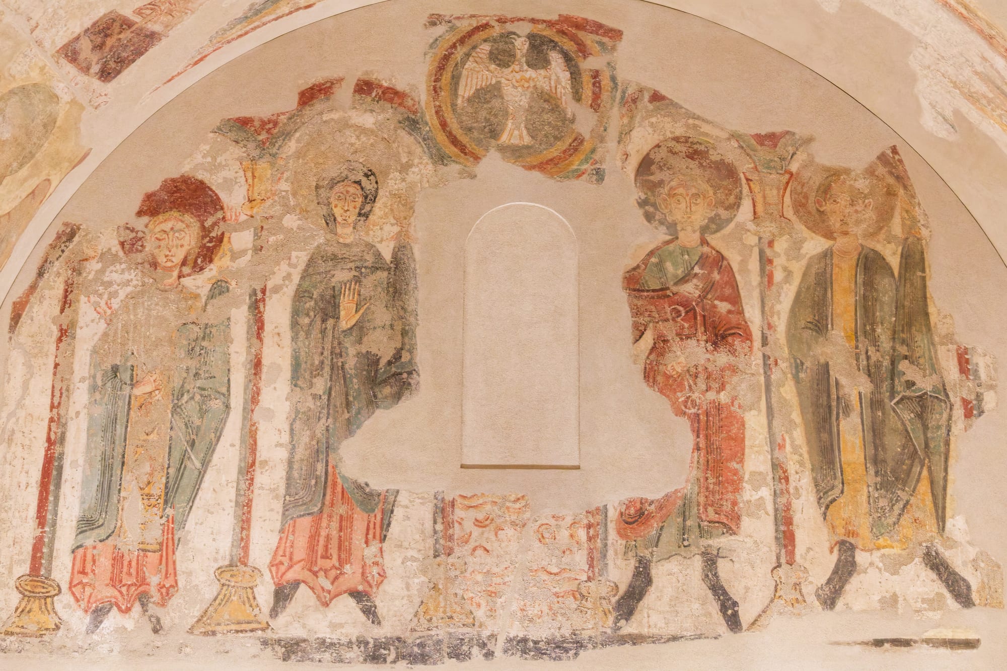 Faded Romanesque wall paintings inside a curved apse showing four standing haloed figures in red, green and ochre robes, with an eagle in a roundel above