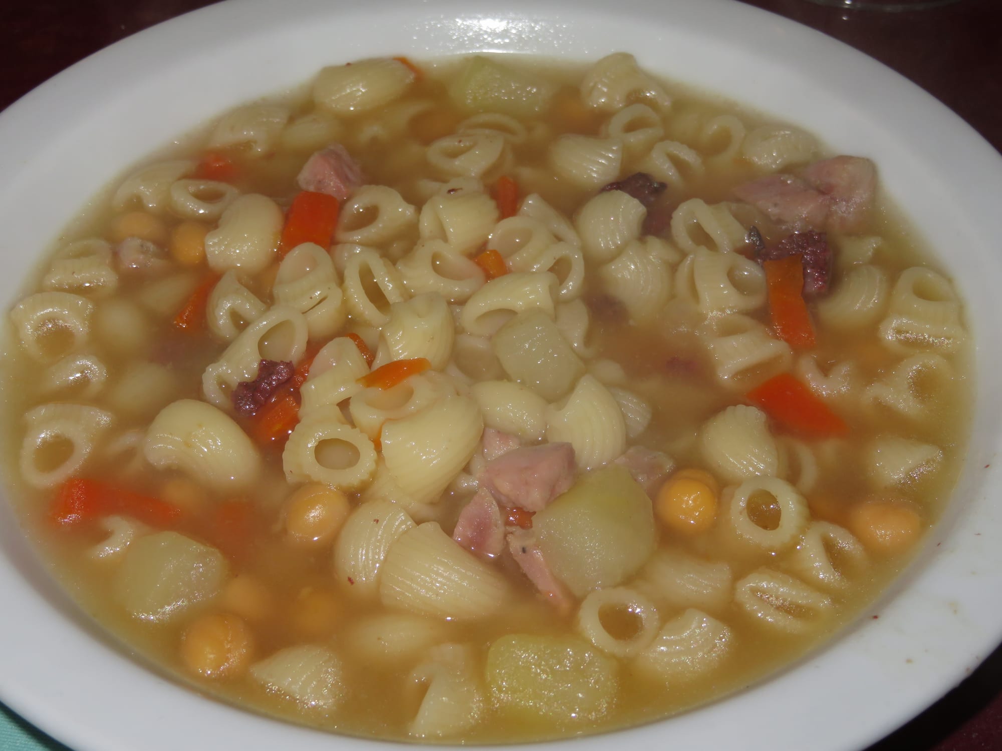 A white bowl of broth filled with small pasta shells, chickpeas, carrot, potato and pieces of ham
